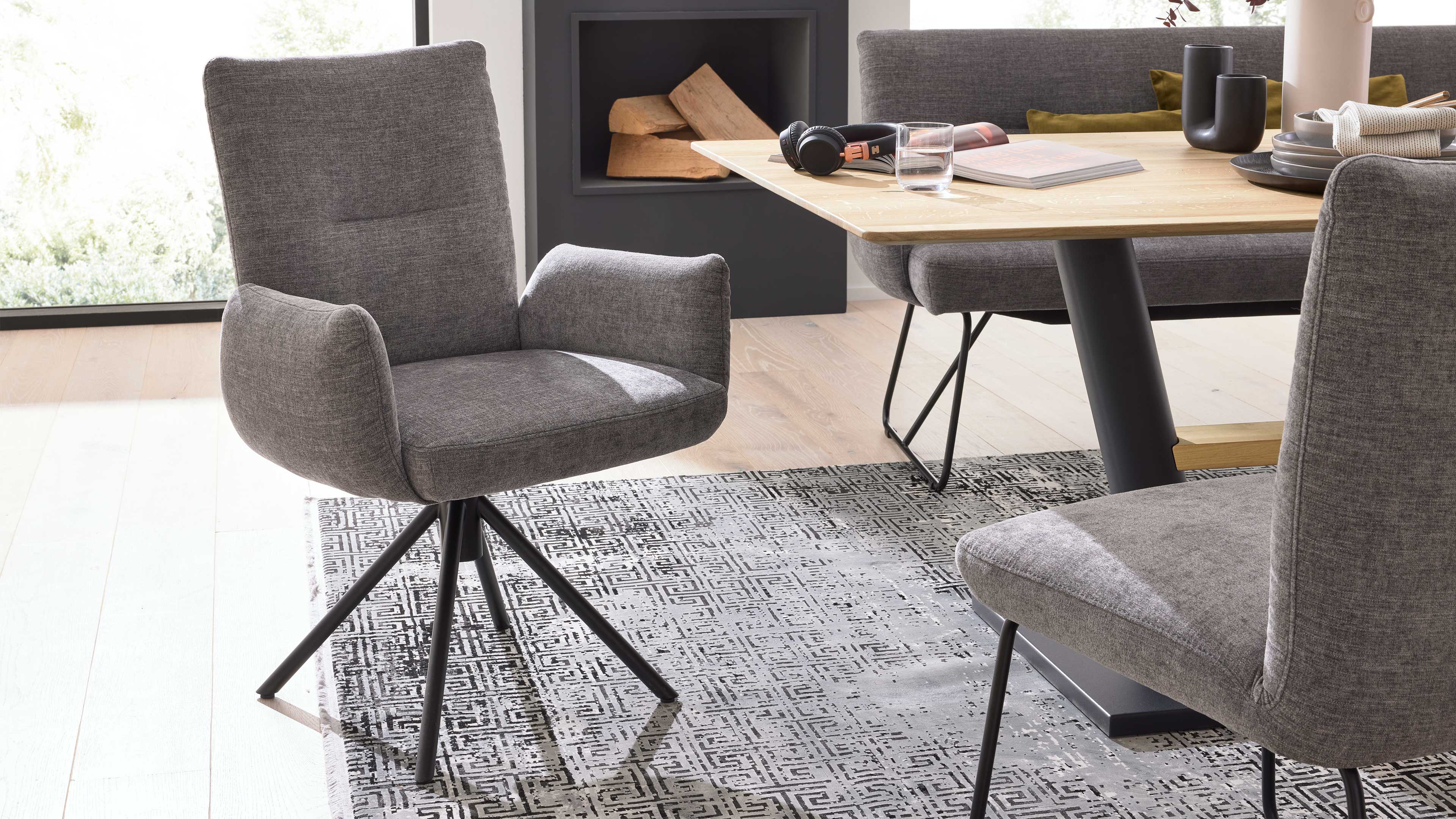 Grauer Polster-Drehstuhl mit niedrigen Armlehnen und schwarzem Sternfuß auf gemustertem Teppich, daneben Esstisch mit Holzplatte.