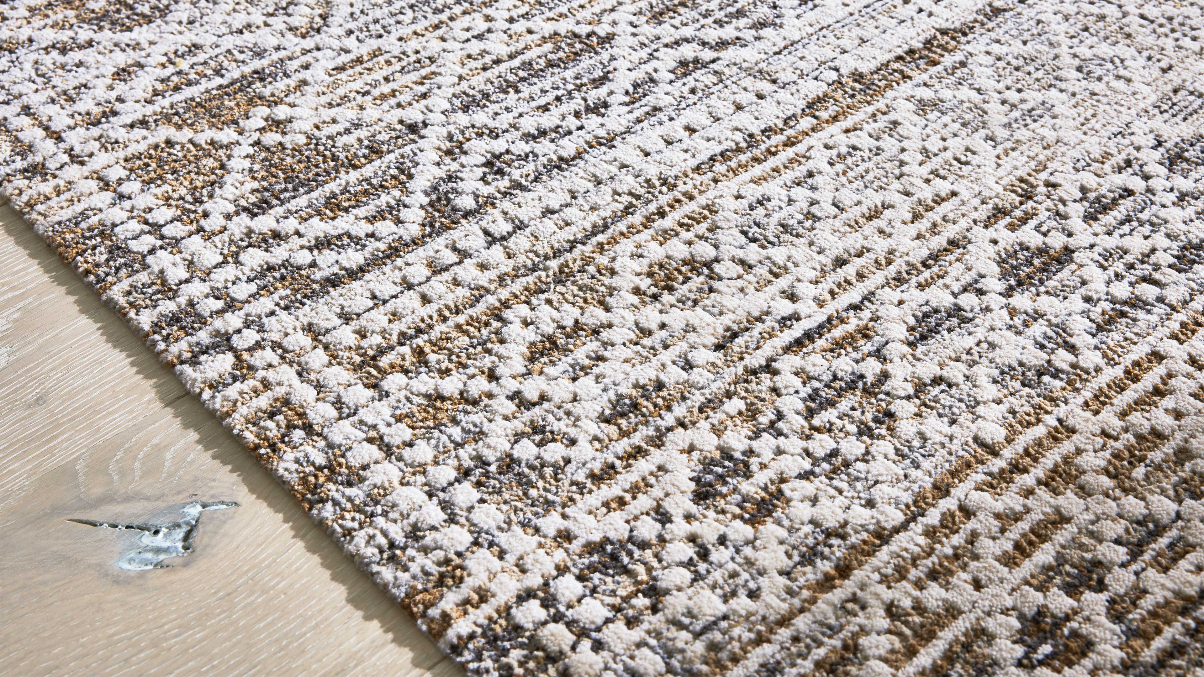 Rechteckiger Teppich mit weiß-beigefarbenem, reliefartigem Punkt-und Linienmuster liegt auf hellem Holzfußboden mit Aststelle.