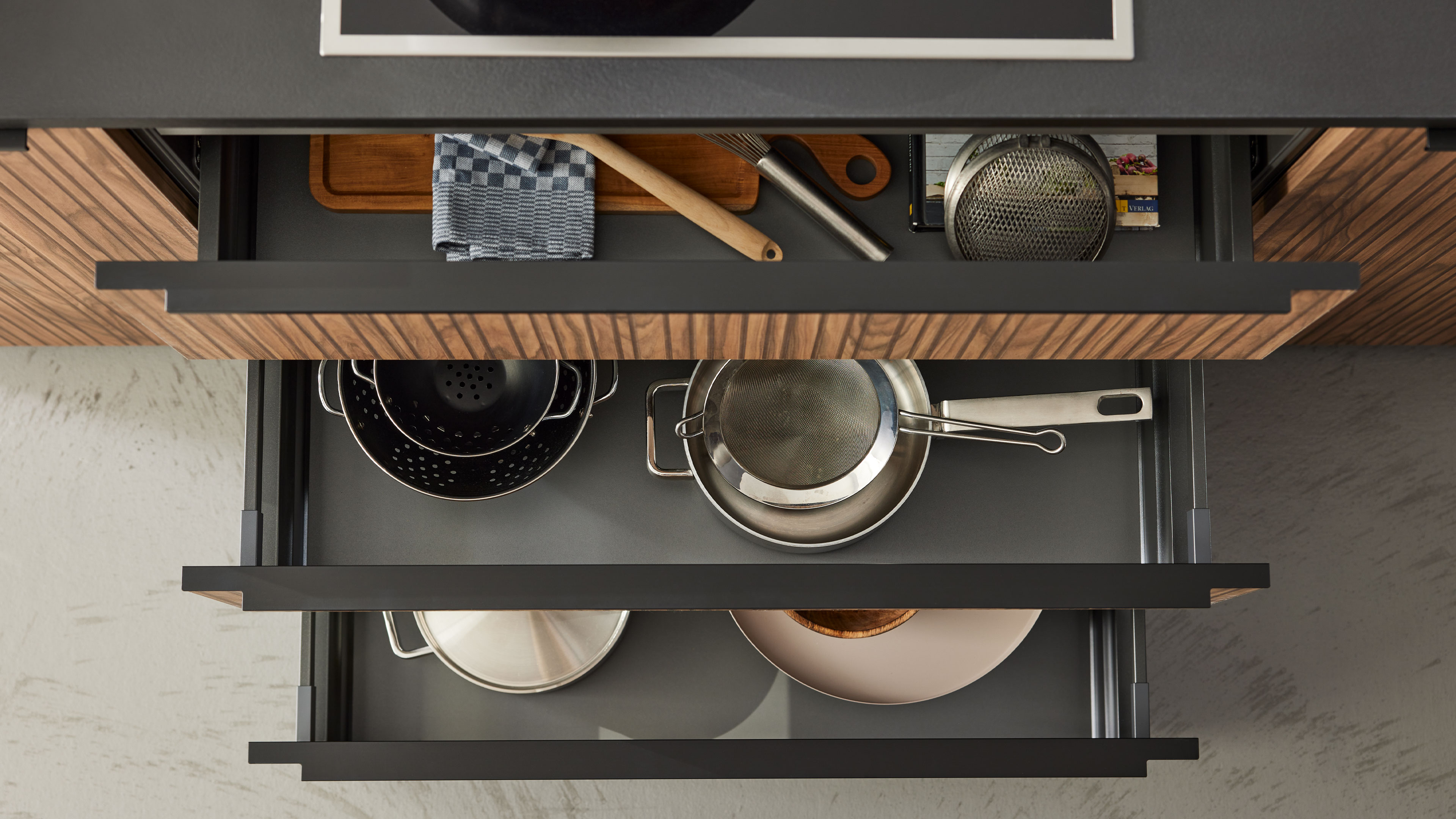 Offene Küchenschubladen mit gerillten Holzfronten, darin ein Schneidebrett und Geschirrtuch, Holzlöffel, Siebe und Töpfe.