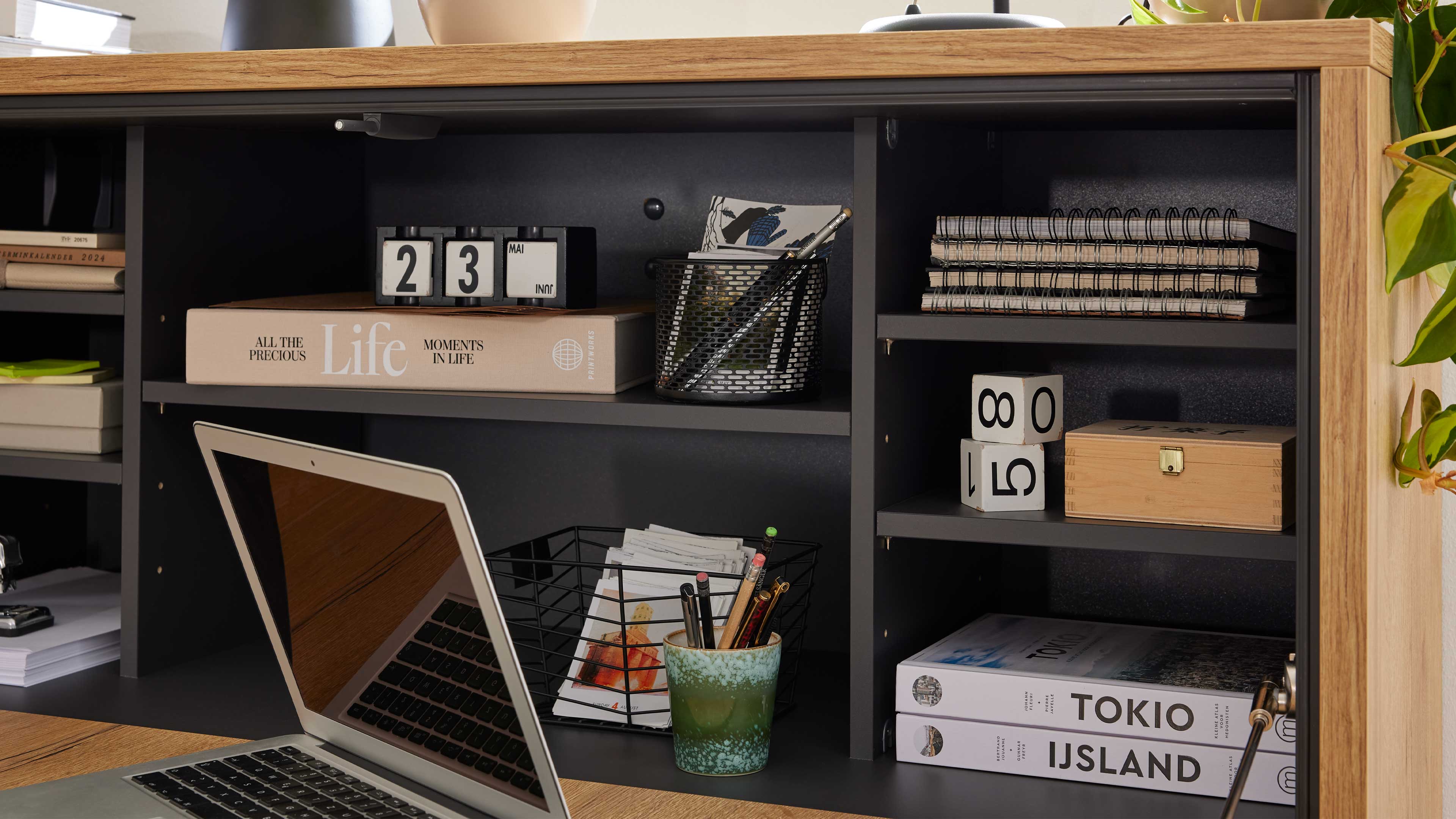 Offenes Schreibtischfach mit Laptop im Vordergrund, Bücherstapel, Kalenderwürfel, Stiftebehälter, Notizblöcken und Holzbox.