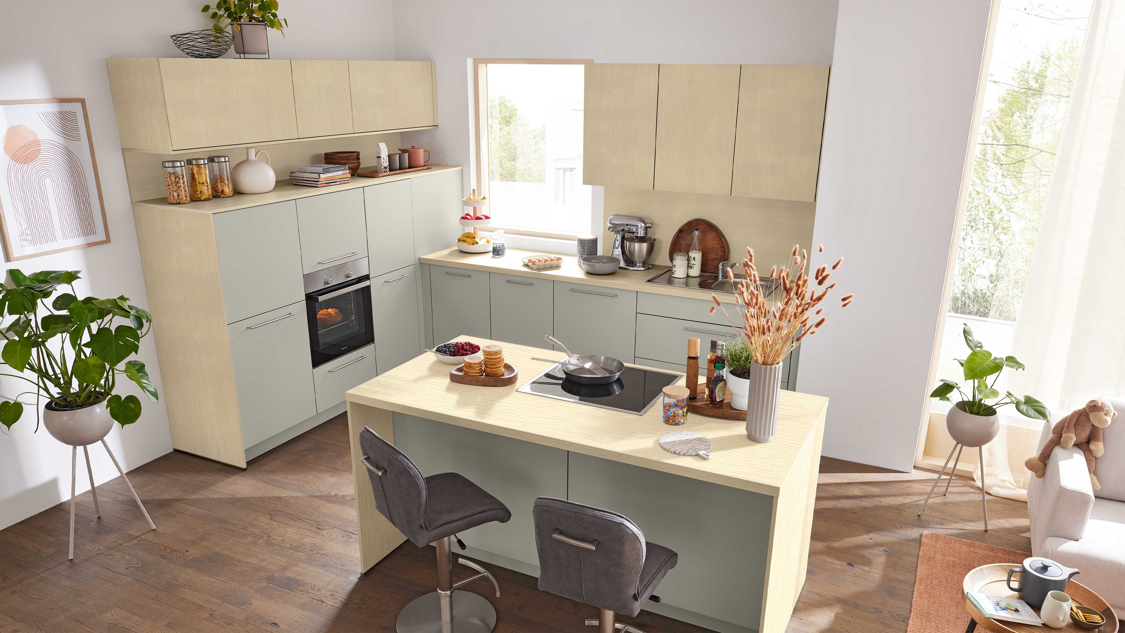 Kücheninsel mit Kochfeld und zwei grauen Barhockern vor hellholz- und salbeigrünen Einbaumöbeln.