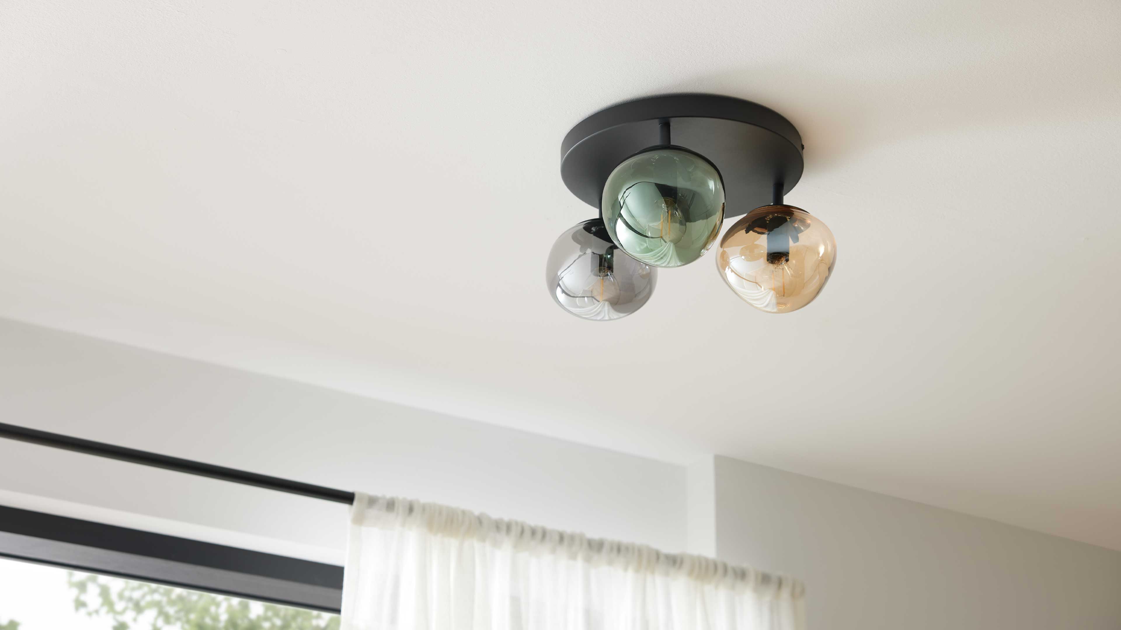 Deckenleuchte mit rundem schwarzen Sockel und drei farbigen Glasglocken (grün, grau, bernstein) über weißer Gardine.