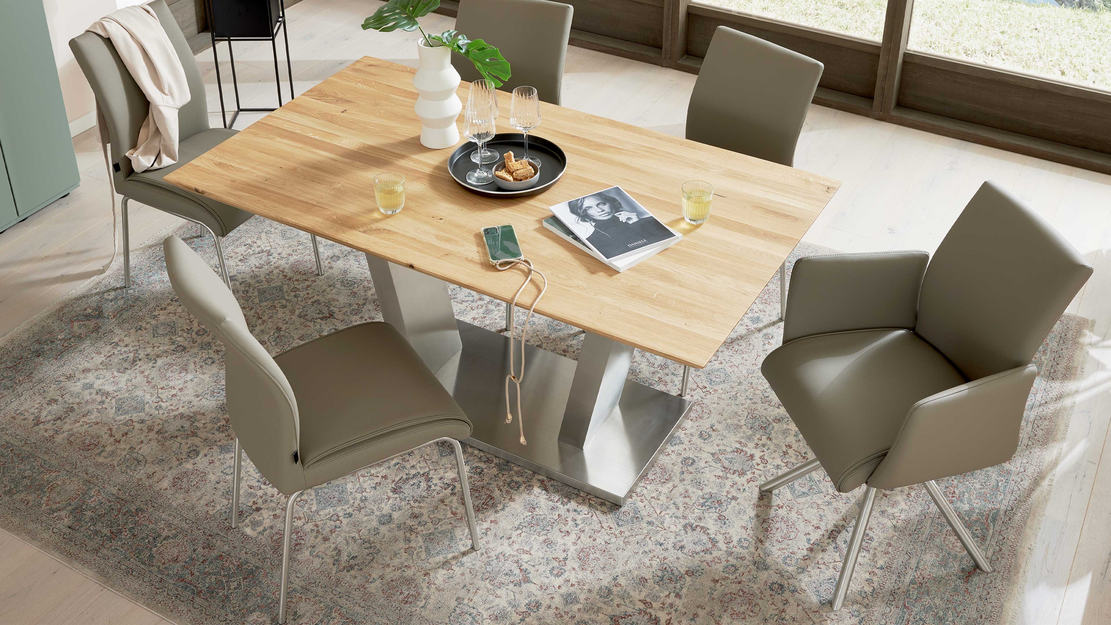 Rechteckiger Esstisch mit heller Holzplatte auf metallischem Doppelständer, umgeben von vier graubraunen gepolsterten Stühlen.