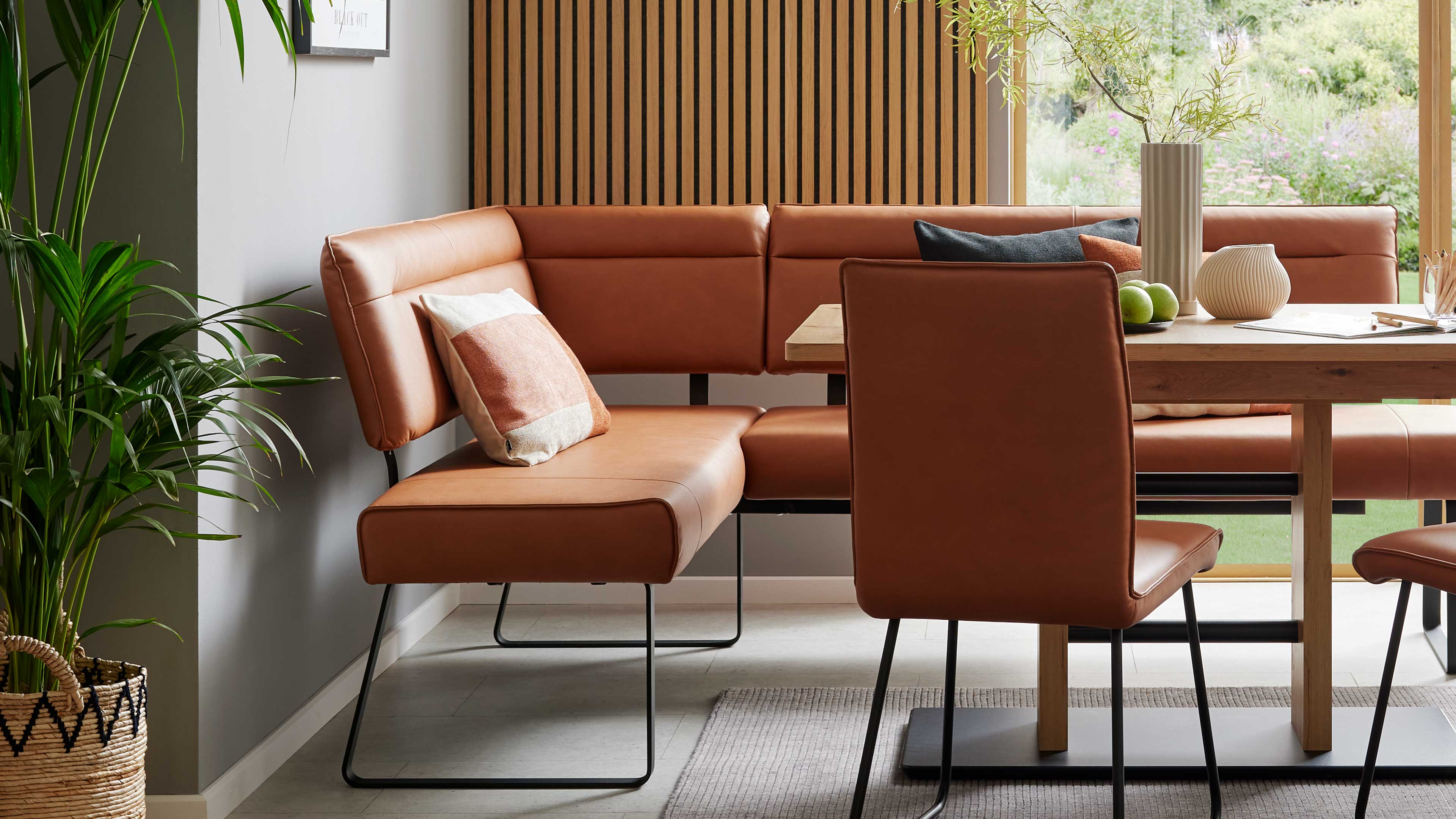 Braun bezogene Eckbank mit schwarzen Metallbeinen an Holztisch, zwei Polsterkissen, Stuhl, Pflanze und Fensterblick.