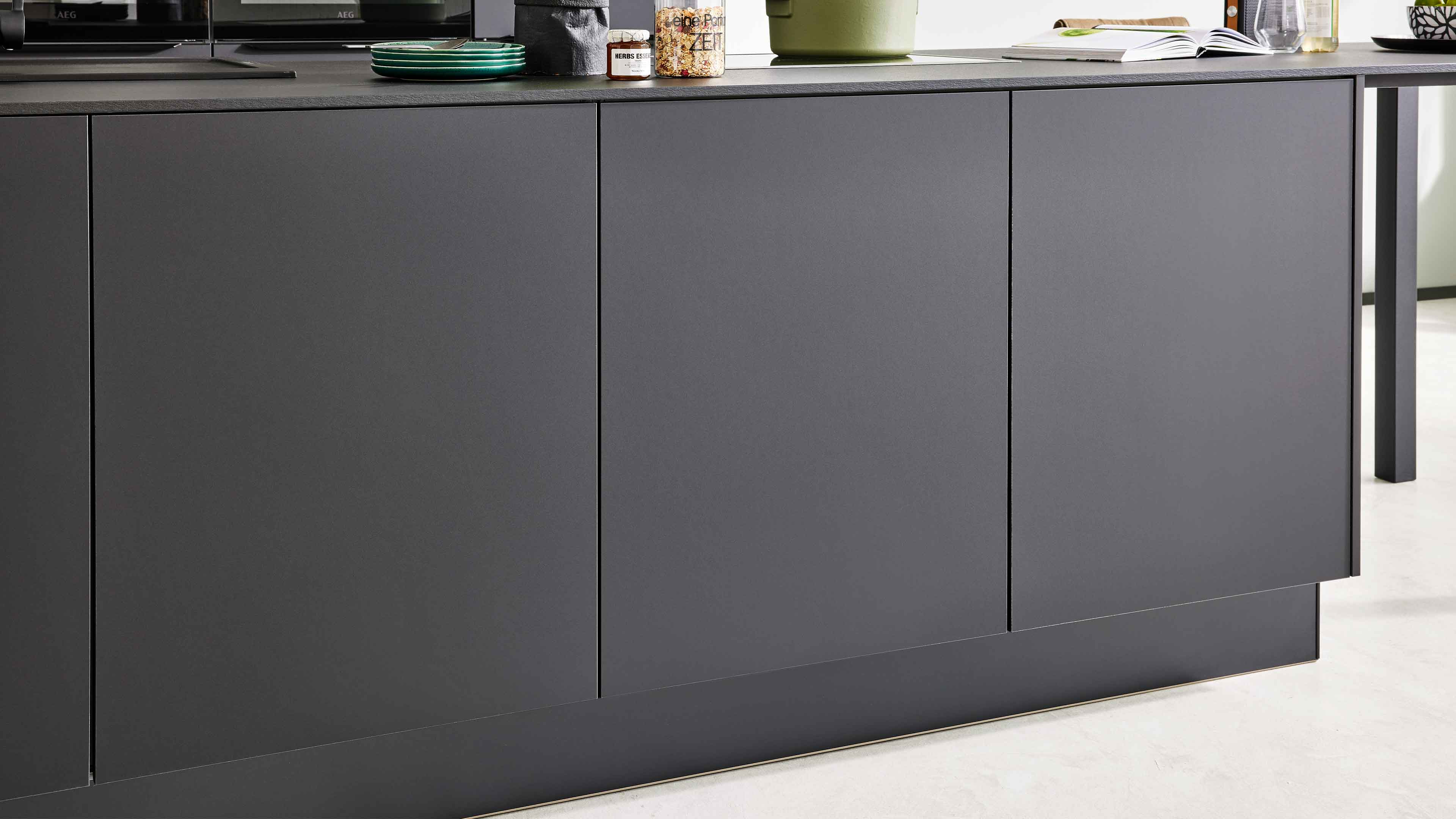 Graue, grifflose Schrankfront mit glatten Türen und schmalen vertikalen Fugen, darauf Teller, Gläser, Dose und Pflanze.