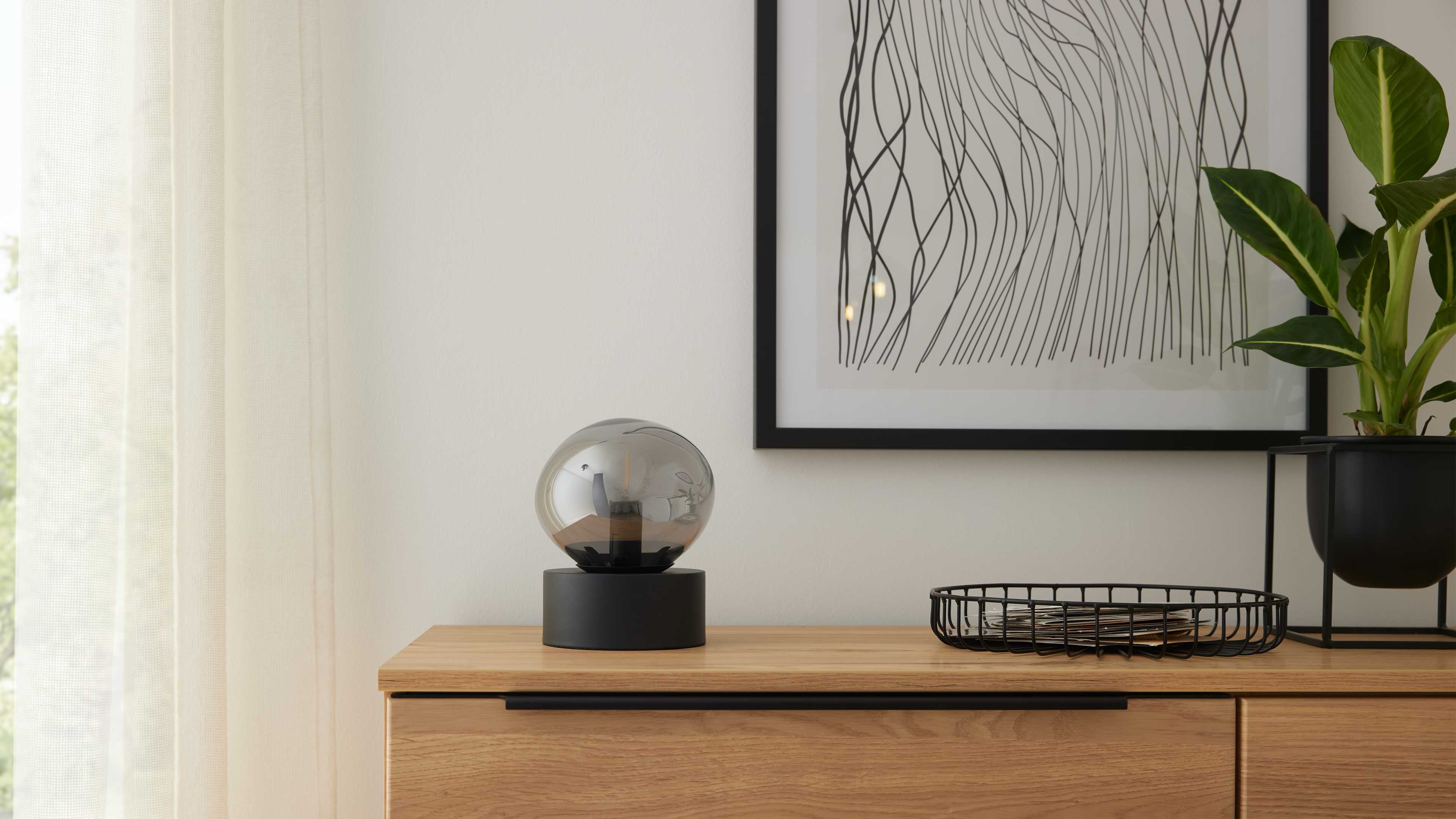 Ein helles Sideboard mit schwarzem Griff, darauf eine rauchglas-Kugellampe auf schwarzem Sockel, ein Drahtkorb und eine Topfpflanze.