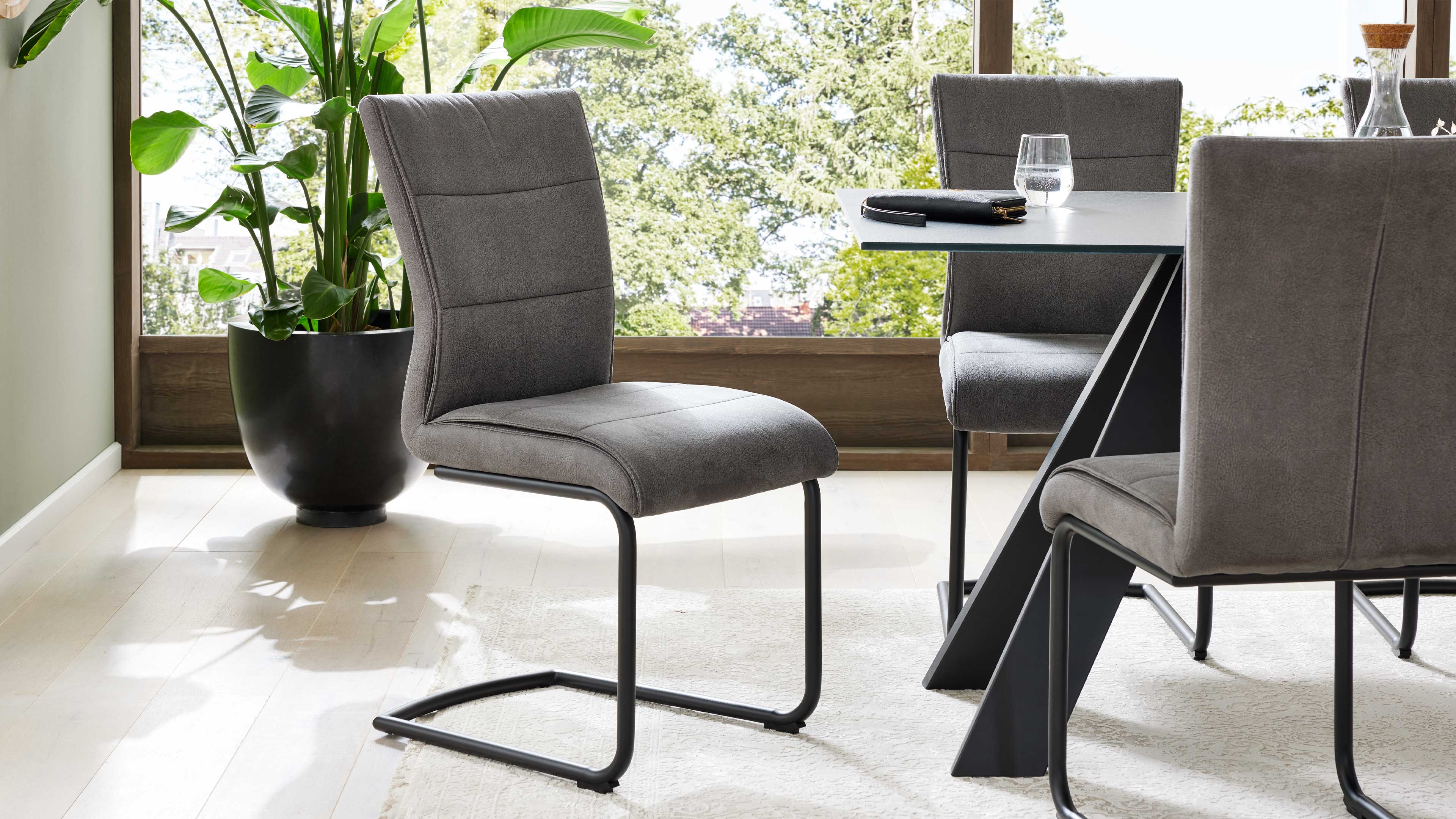 Grau gepolsterter Freischwinger mit schwarzem Metallgestell am Esstisch, auf dem Tisch Glas und dunkler Gegenstand, links Topfpflanze.