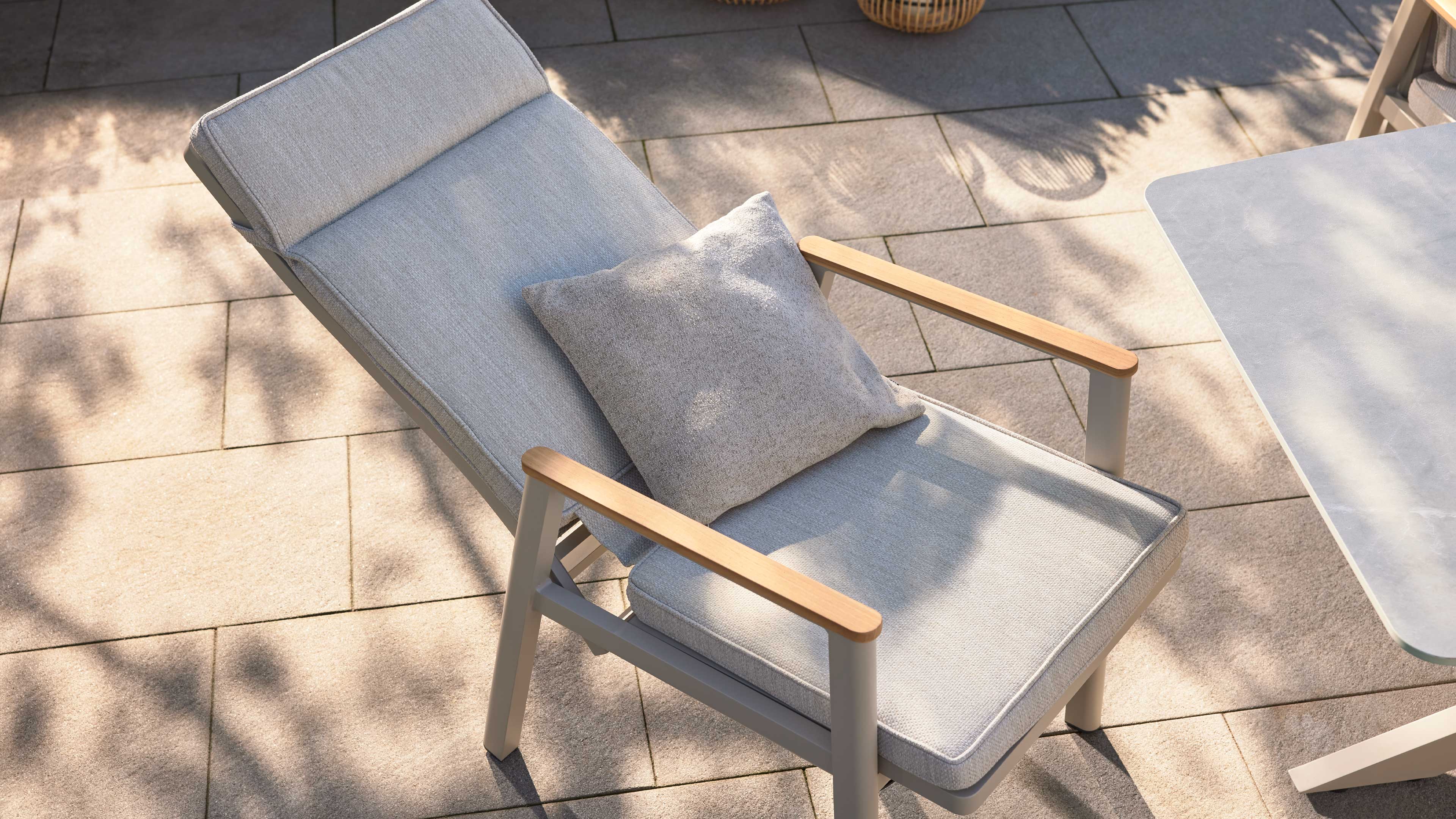 Graue gepolsterte Gartenliege mit quadratischem Rückenkissen und Holzarmlehnen auf Terrassenplatten, mit Pflanzenschatten.