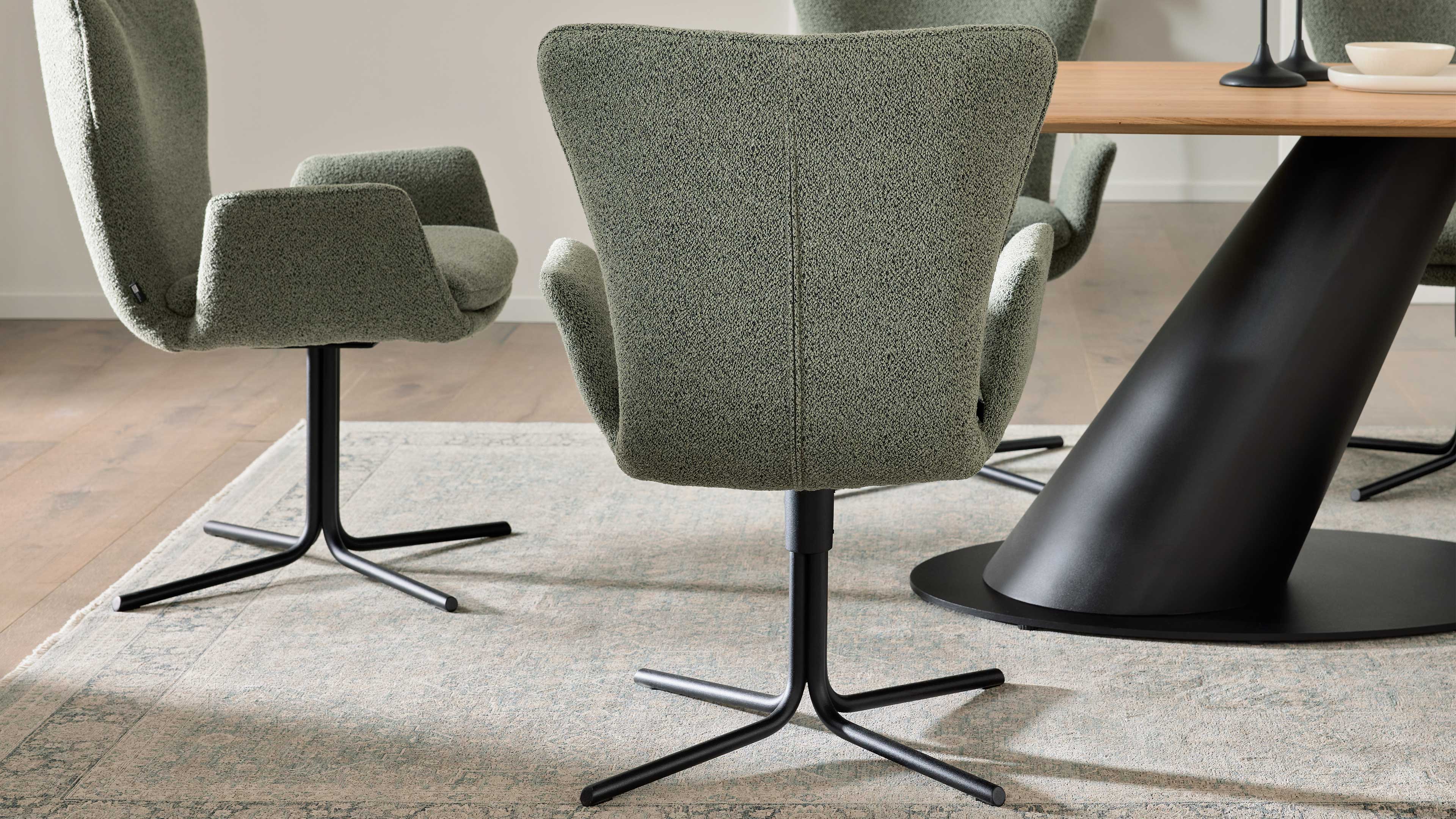 Grüner Drehstuhl mit schwarzem Viersternfuß im Vordergrund, dahinter weitere Stühle an Holztisch mit schwarzem, schrägem Fuß auf Teppich.