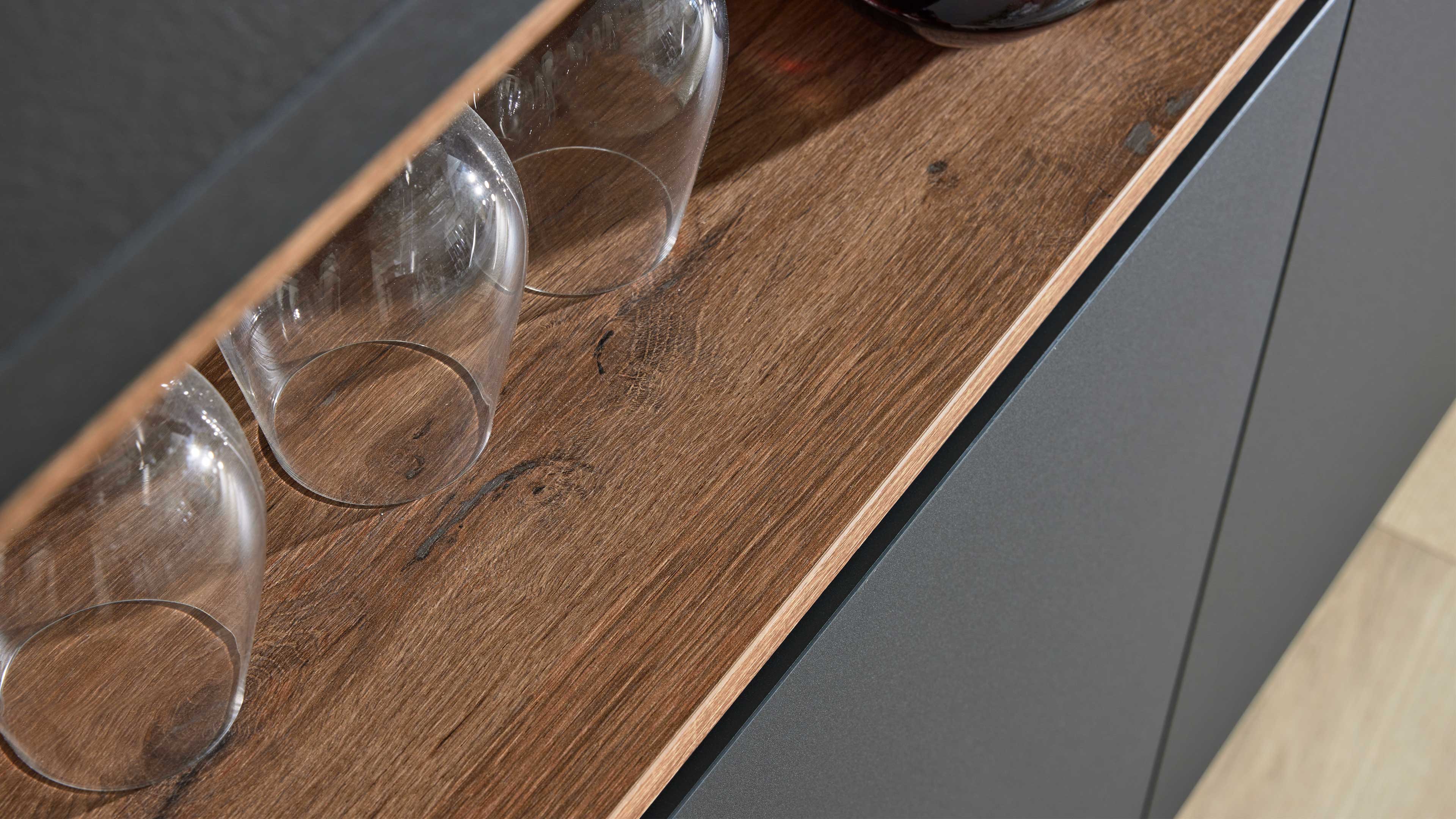 Holzplatte mit sichtbarer Maserung, drei umgedrehte Gläser darauf und dunkelgraue Schrankfronten unterhalb.