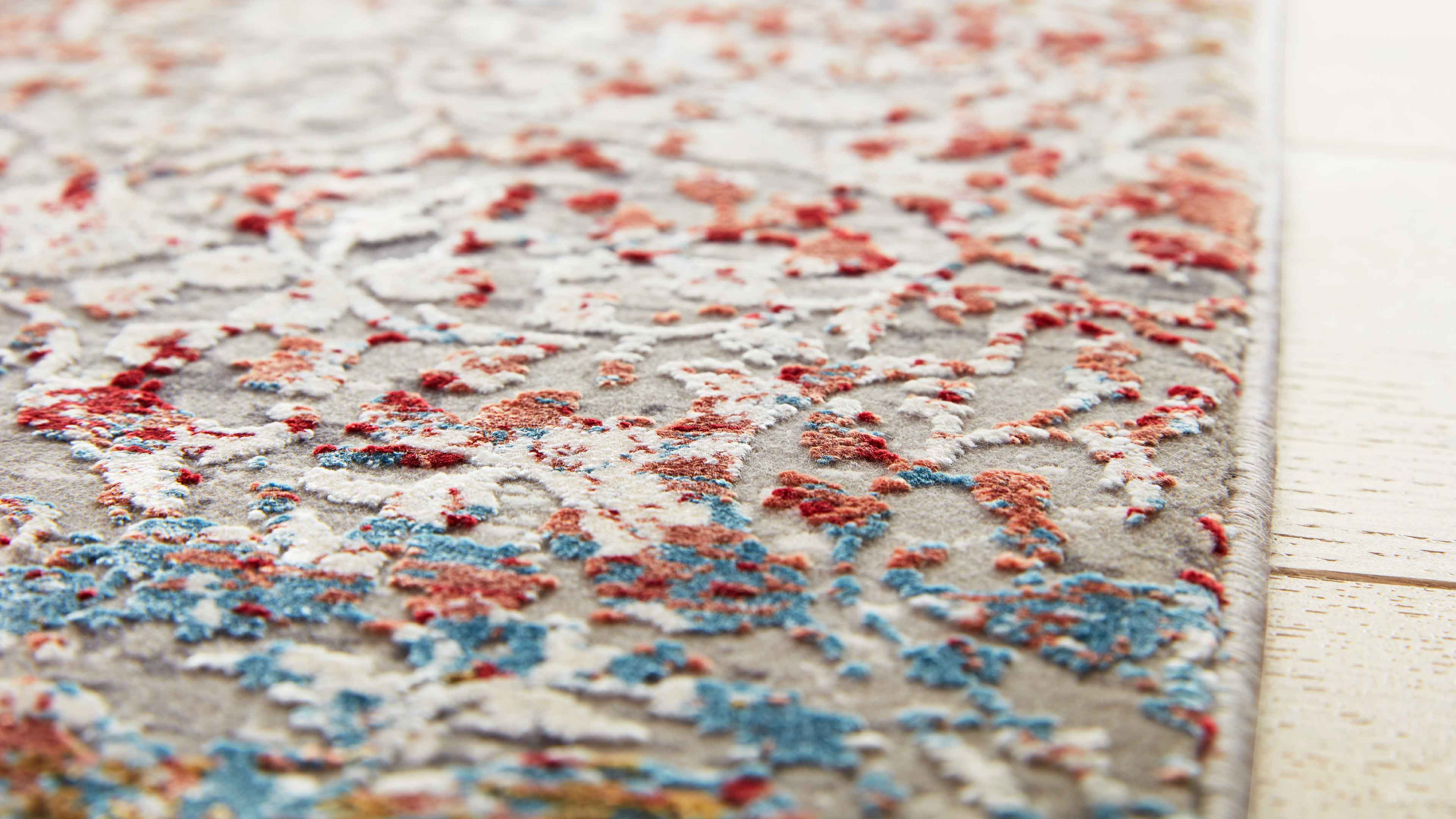 Nahaufnahme eines Teppichs mit erhabener Musterstruktur in Rot, Blau und Weiß auf hellem Grund, rechts Holzfußboden.