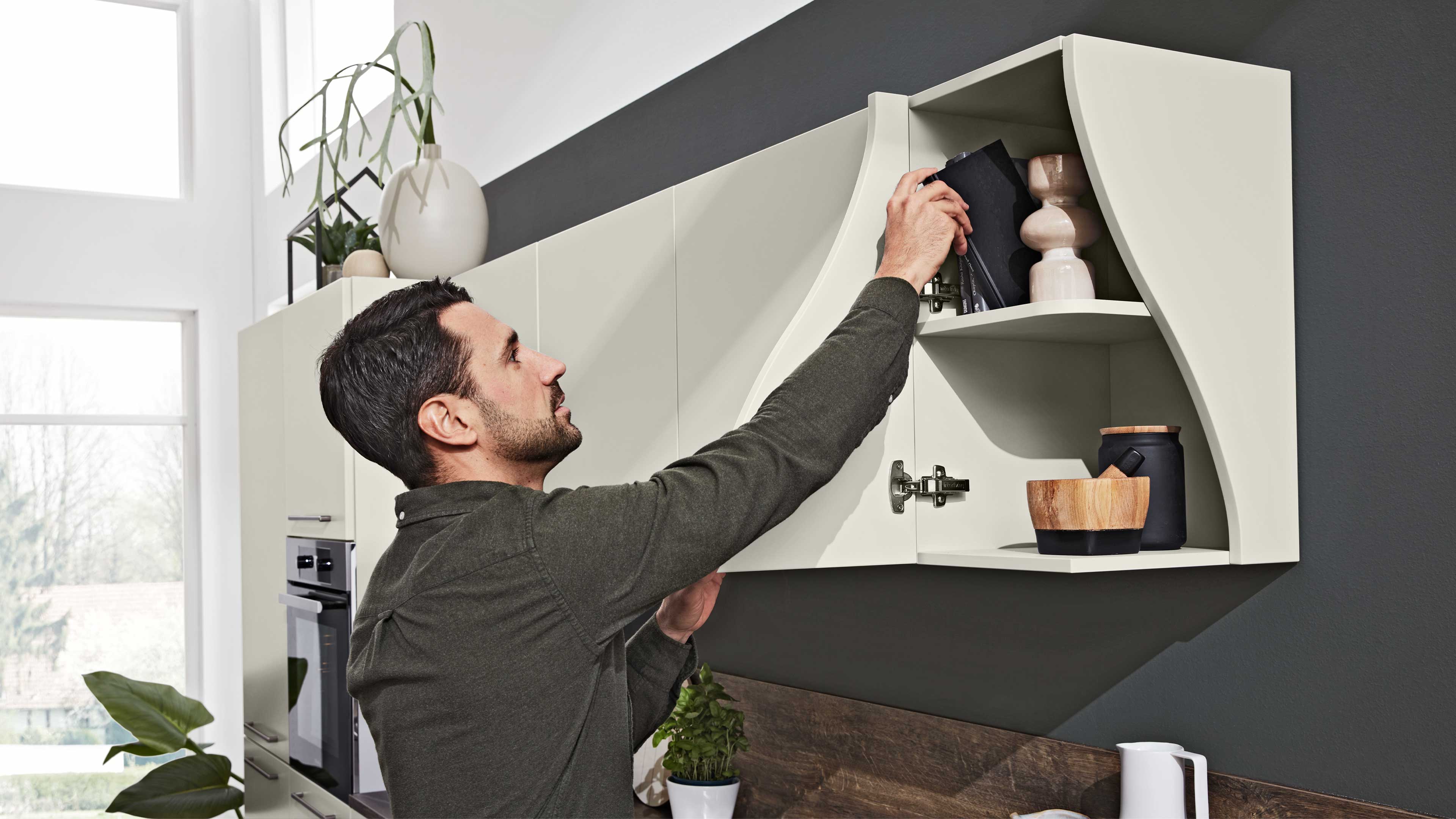 Ein Mann greift in ein helles, wandhängendes Küchenschrankfach mit geschwungenen Seiten, darin Bücher, Keramikgefäße und Holzschale.