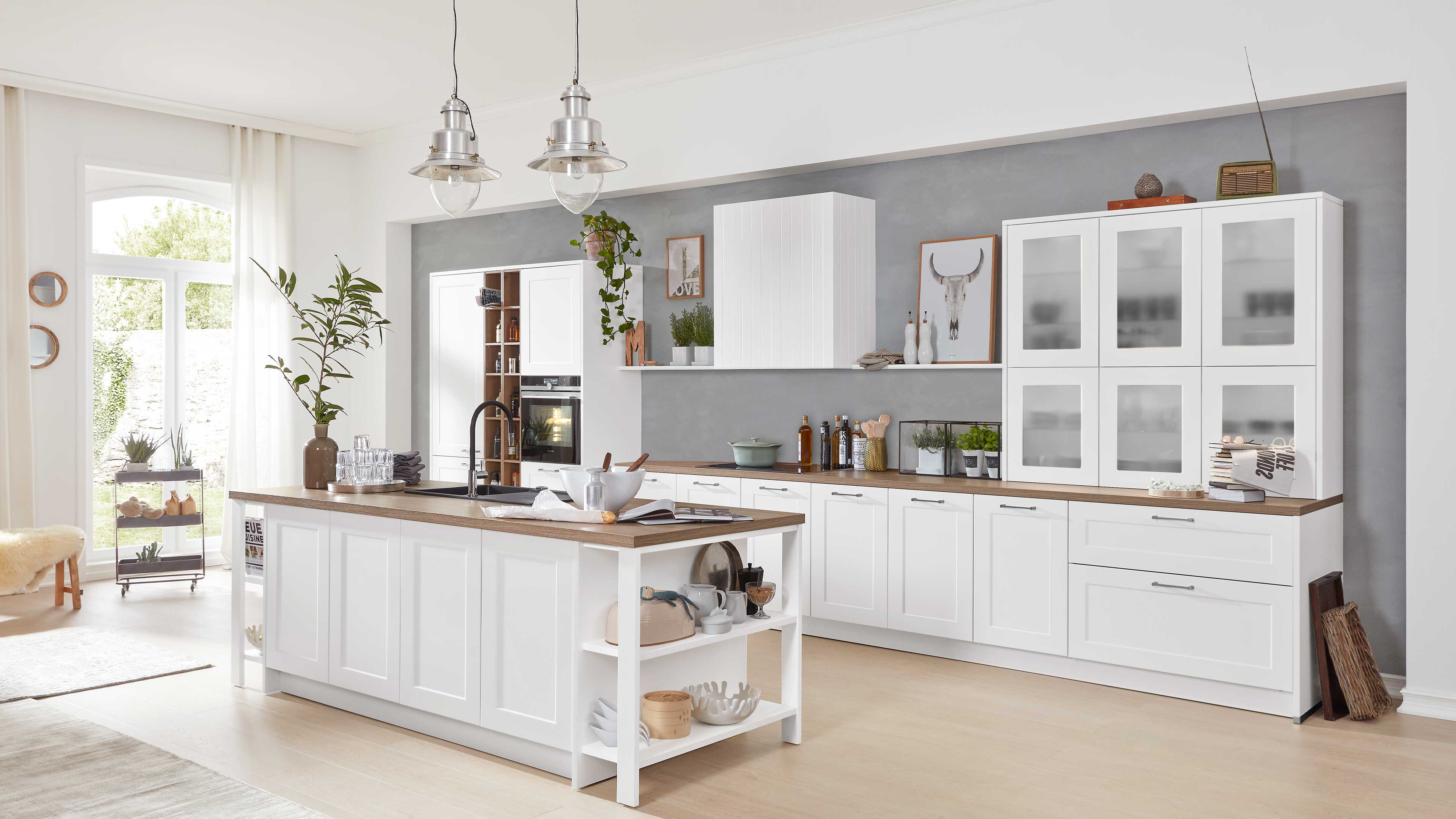 Helle Einbauküche mit weißer Kücheninsel, holzfarbener Arbeitsplatte, integrierter Spüle, Glasvitrinen und metallischen Hängelampen.