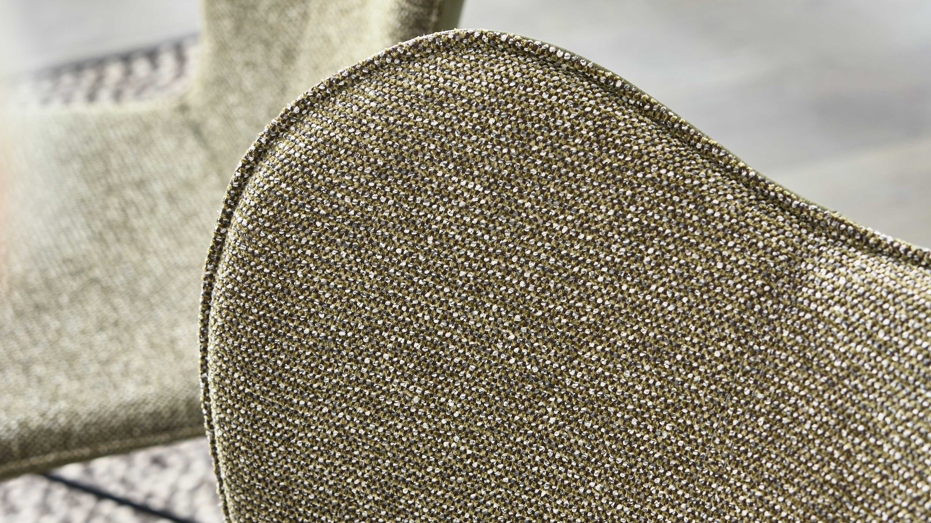 Nahaufnahme der abgerundeten Rückenlehne eines Stuhls mit olivefarbenem, gewebtem Stoff und sichtbarer Kappnaht, dahinter unscharfer Sitz.