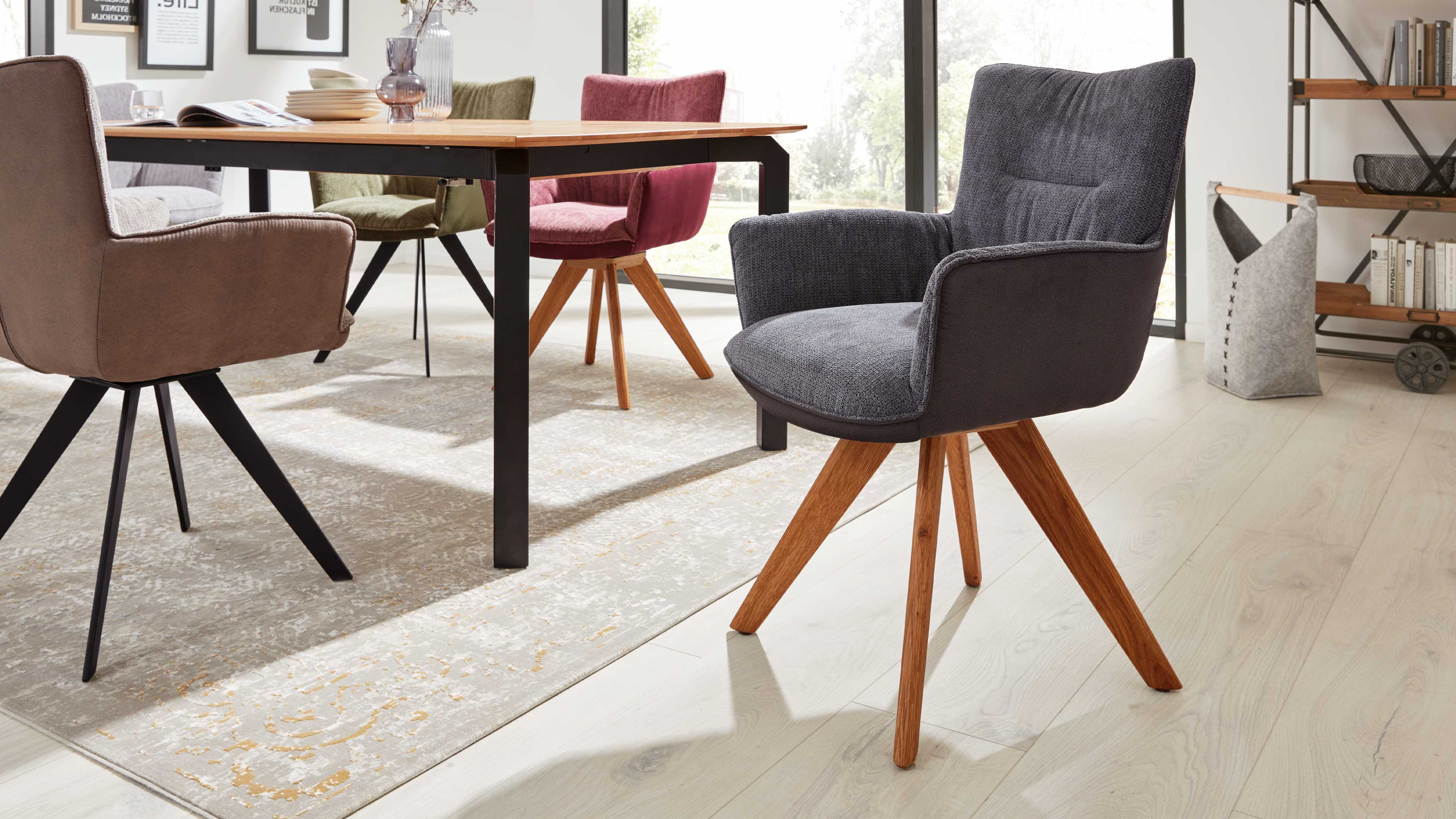 Dunkelgrauer, gepolsterter Armlehnstuhl mit schräggestellten Holzbeinen im Vordergrund vor Esstisch und weiteren Stühlen, Teppich und Regal.