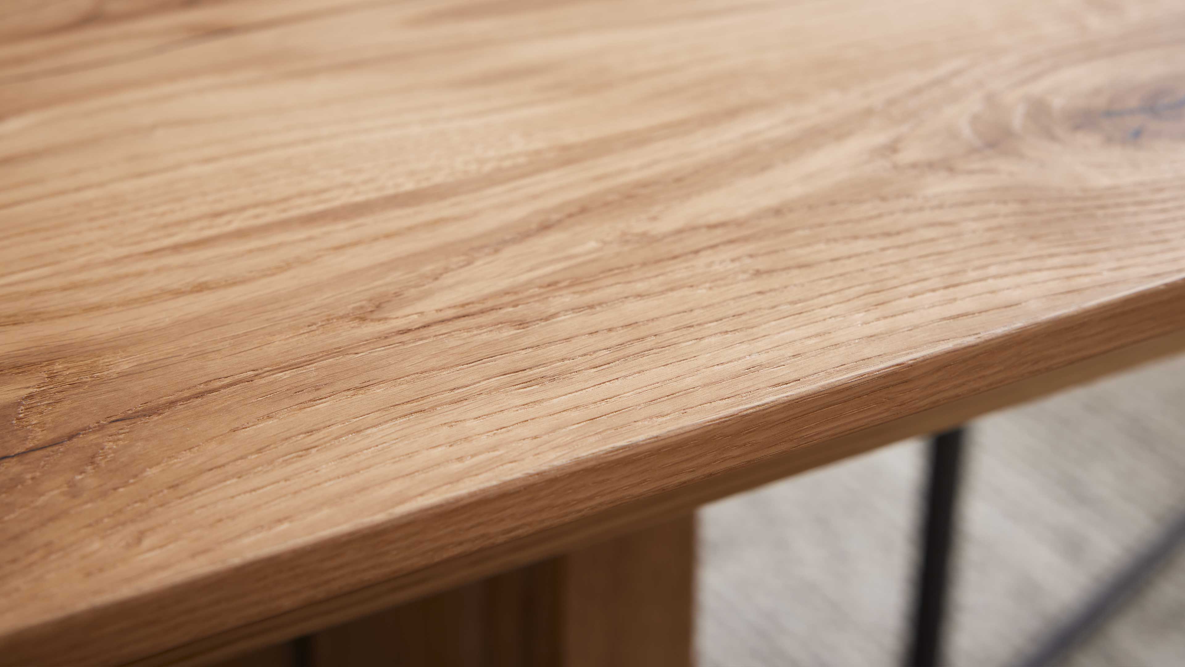 Nahaufnahme einer hellen Holzplatte mit sichtbarer Maserung und abgerundeter Kante, darunter schwarzes Metallbein.