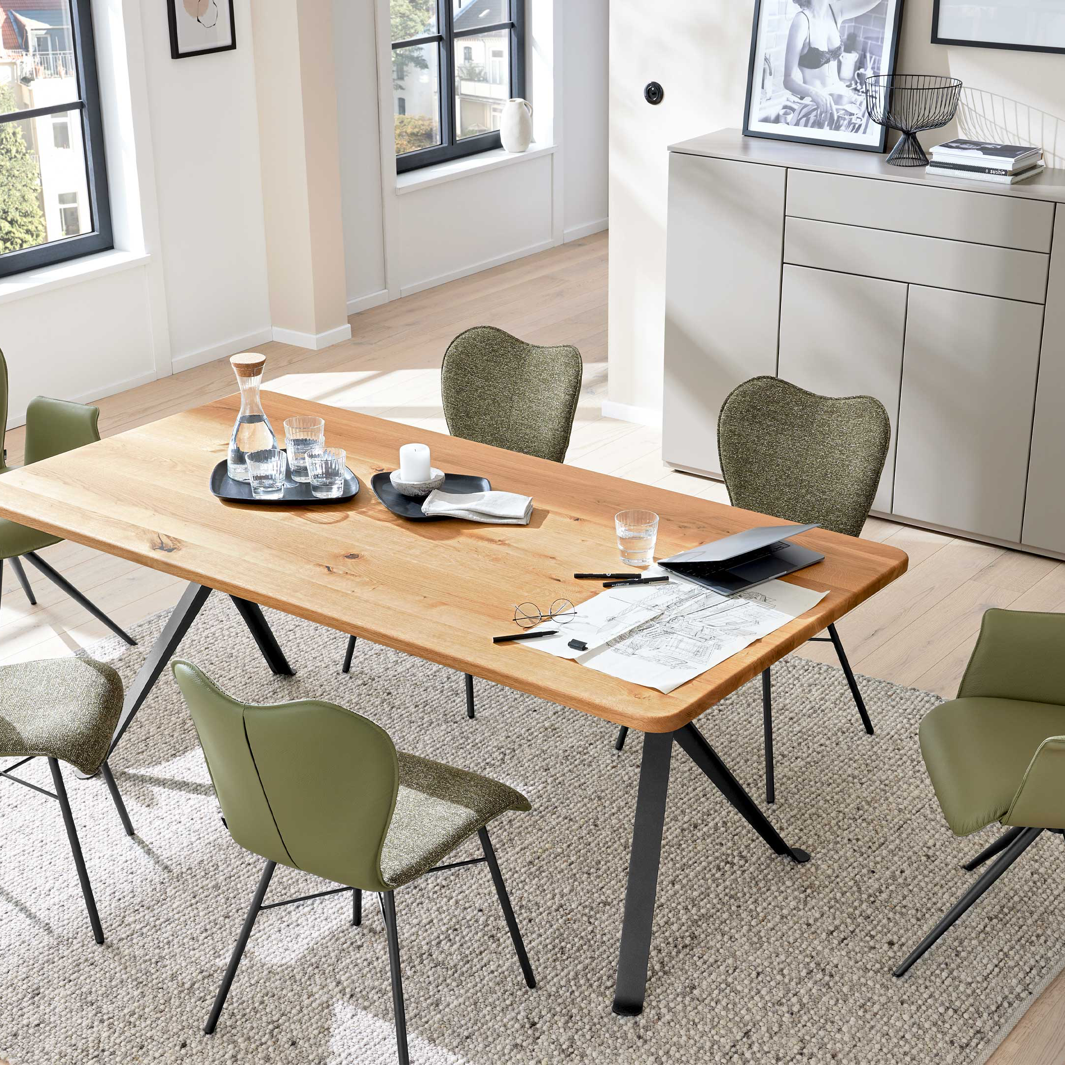 Esstisch aus hellem Holz mit schwarzen Metallbeinen, umgeben von sechs grünen Stühlen auf Teppich, Sideboard und Pflanzen.