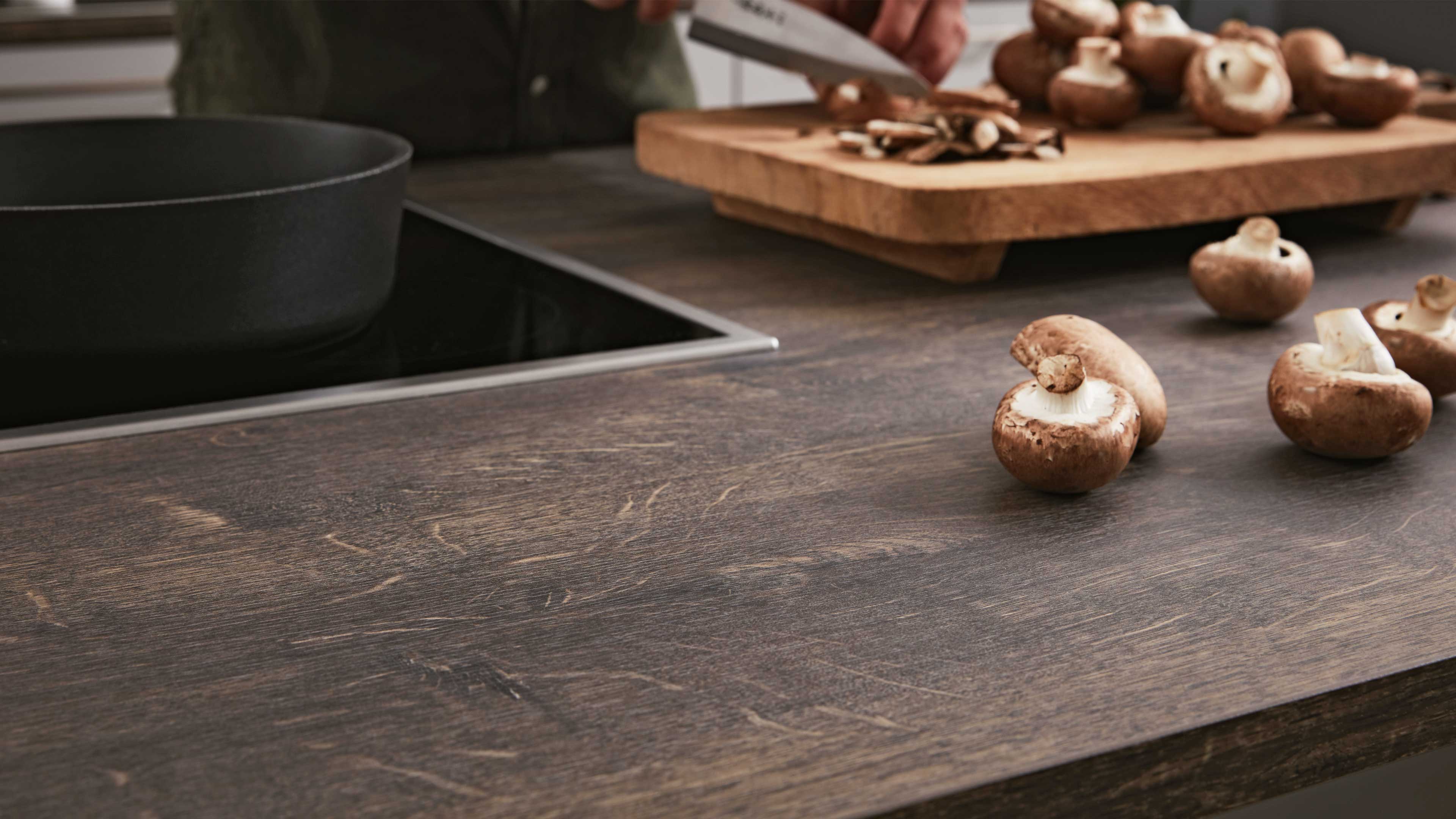 Dunkle Holzarbeitsplatte mit verstreuten braunen Champignons, rechts ein Schneidebrett und Hand beim Schneiden.