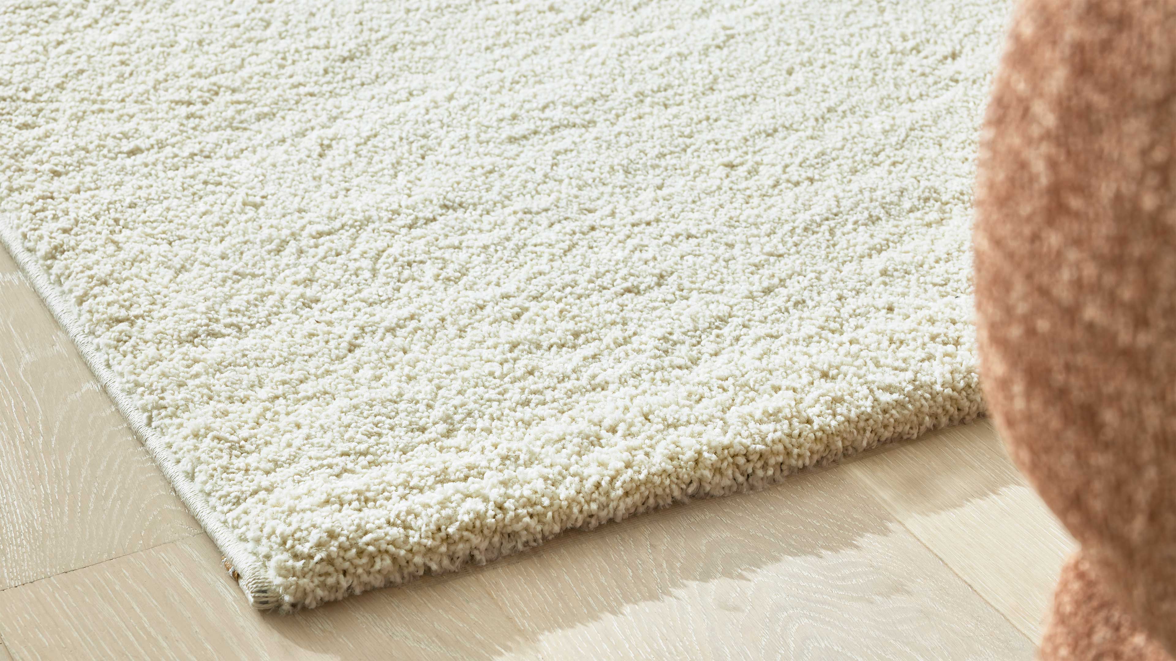 Cremefarbener, dichter Schlingen-Teppich liegt an der Kante auf hellem Holzfußboden, rechts teilweise brauner Stoff eines Sitzmöbels.