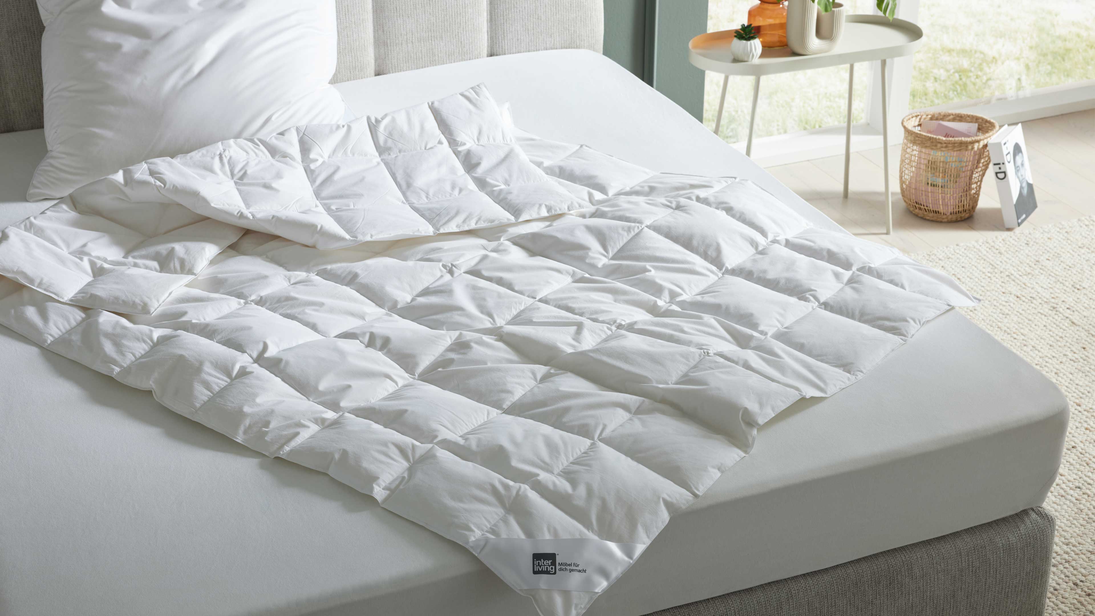 Weiße gesteppte Bettdecke auf hellgrauer Matratze und grauem Bettrahmen, links Kissen, rechts Beistelltisch mit Vasen und Korb.