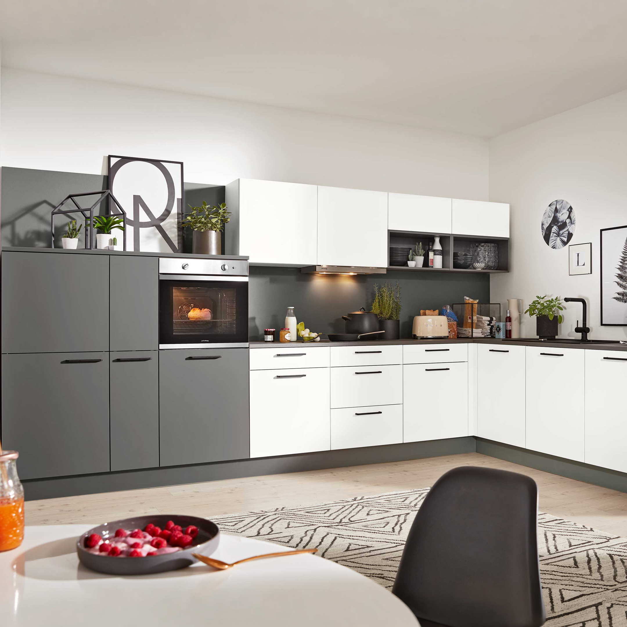 Eckküche mit weißen Hängeschränken und grauen Unterschränken, eingebautem Ofen, schwarzer Arbeitsplatte, Pflanzen und hängender Lampe.