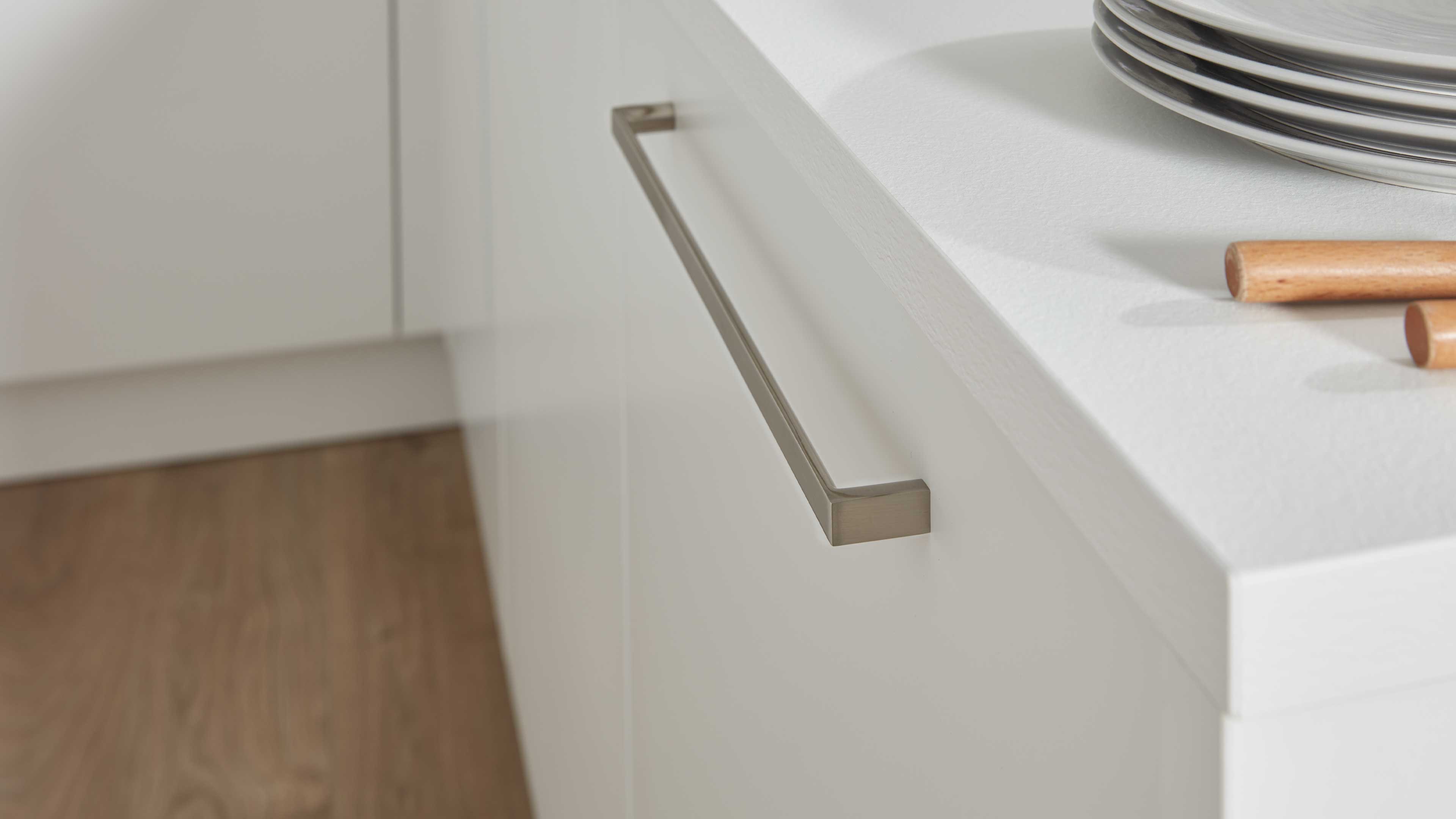 Weiße Küchenschublade mit langem rechteckigem Metallgriff in gebürsteter Optik, weiße Arbeitsplatte, Holzstiele und Tellerrand sichtbar.
