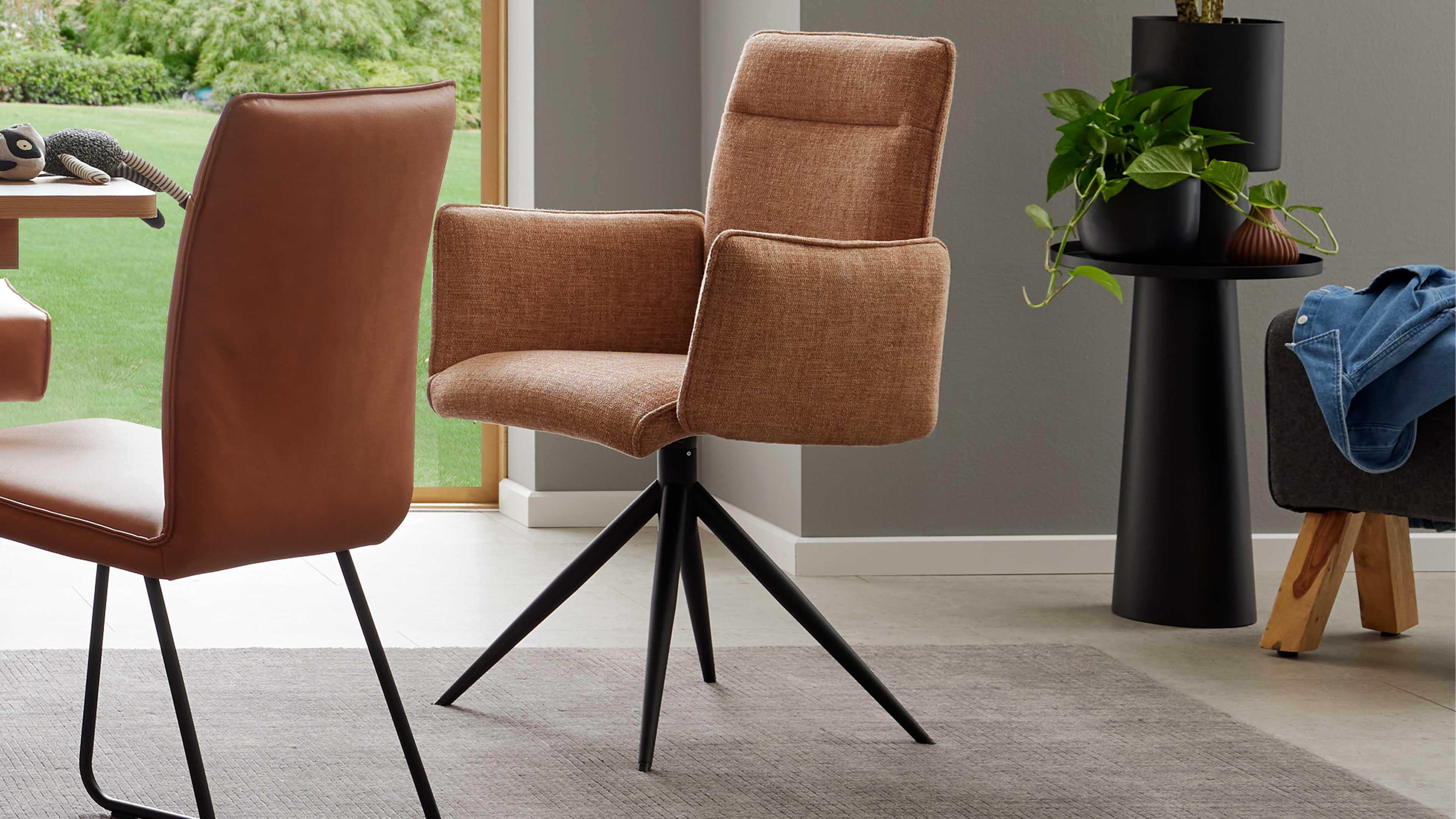 Brauner Drehstuhl aus Stoff mit gepolsterten Armlehnen und schwarzem vierarmigem Metallfuß, links Ledersitz, rechts schwarzer Beistelltisch.