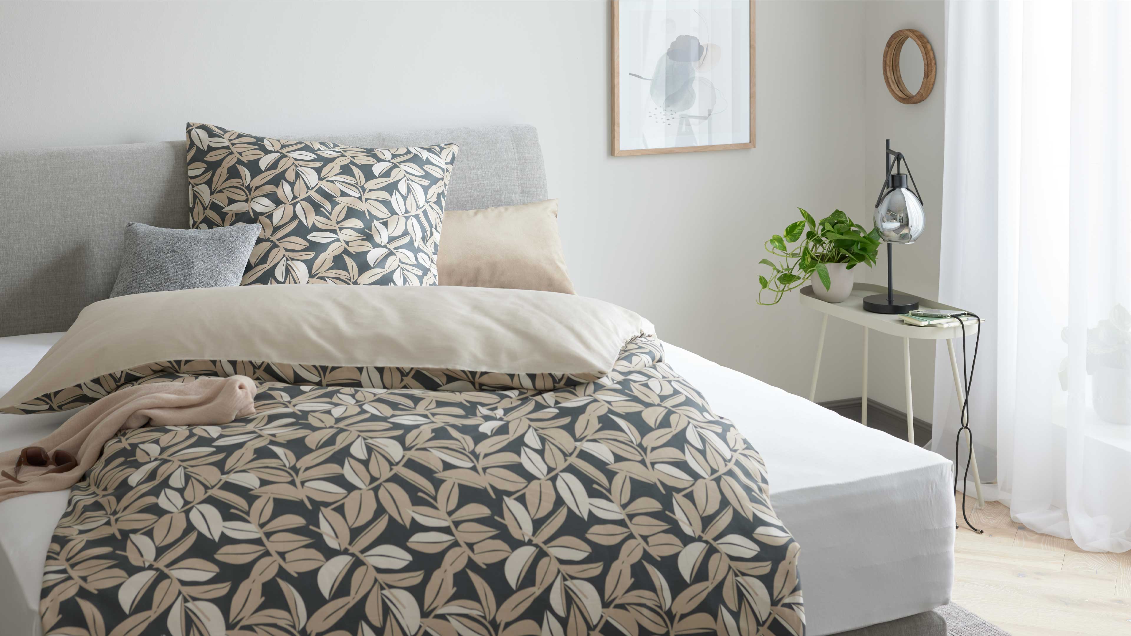 Bett mit hellgrauem Kopfteil und beige-dunkel gemustertem Blatt-Bettbezug, daneben Beistelltisch mit Lampe und Topfpflanze.