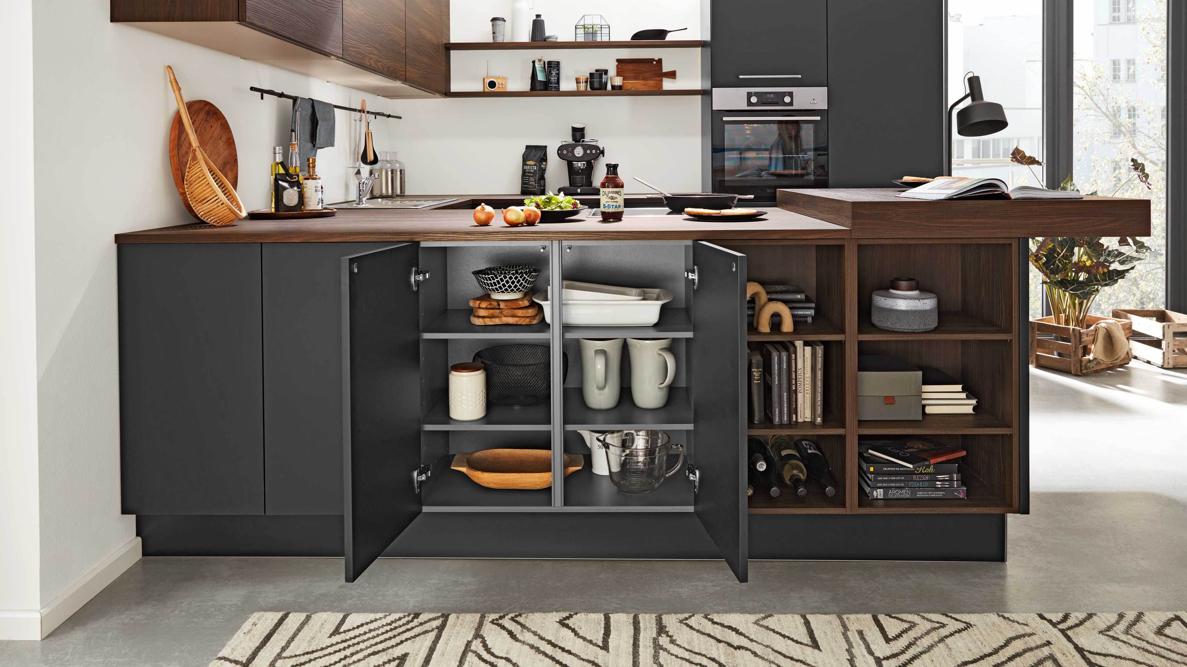 Dunkelgrauer Unterschrank mit offenen Türen, Schüsseln, Tassen und Backformen; rechts offenes Holzregal mit Büchern und Wein.