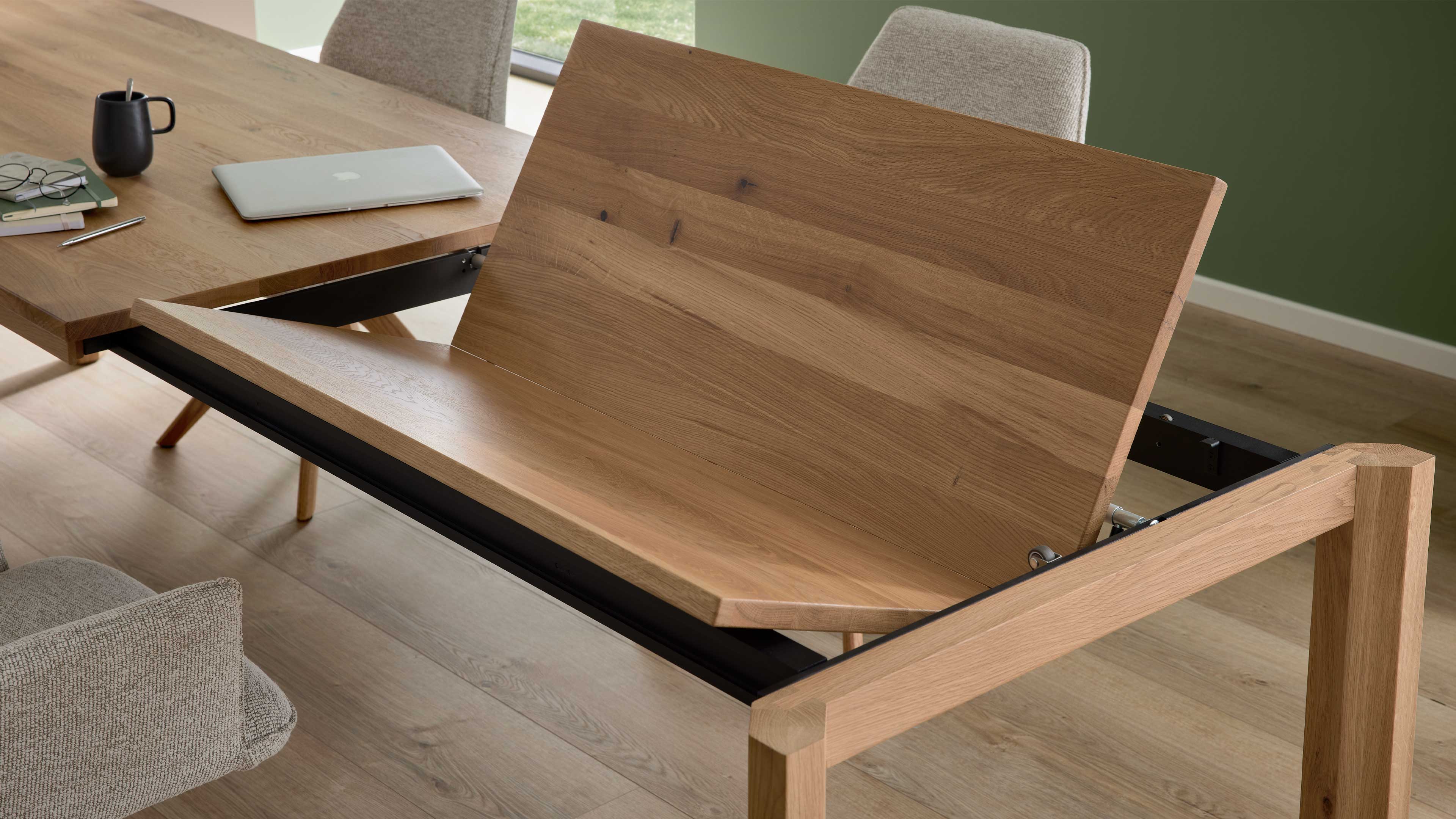 Ausgezogener hölzerner Esstisch mit aufgeklappter Verlängerungsplatte in schwarzer Metallführung, beige Polsterstühle und Laptop.