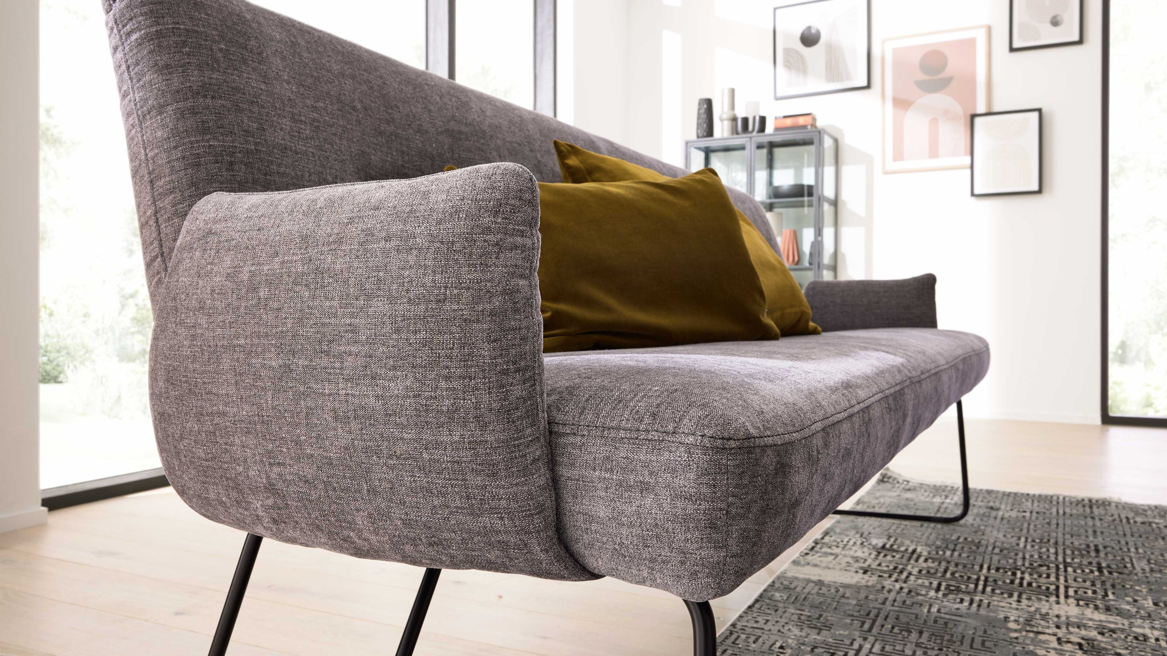 Graues, gepolstertes Sofa mit schlanken schwarzen Metallbeinen und zwei senfgelben Kissen, davor Teppich und Vitrine.