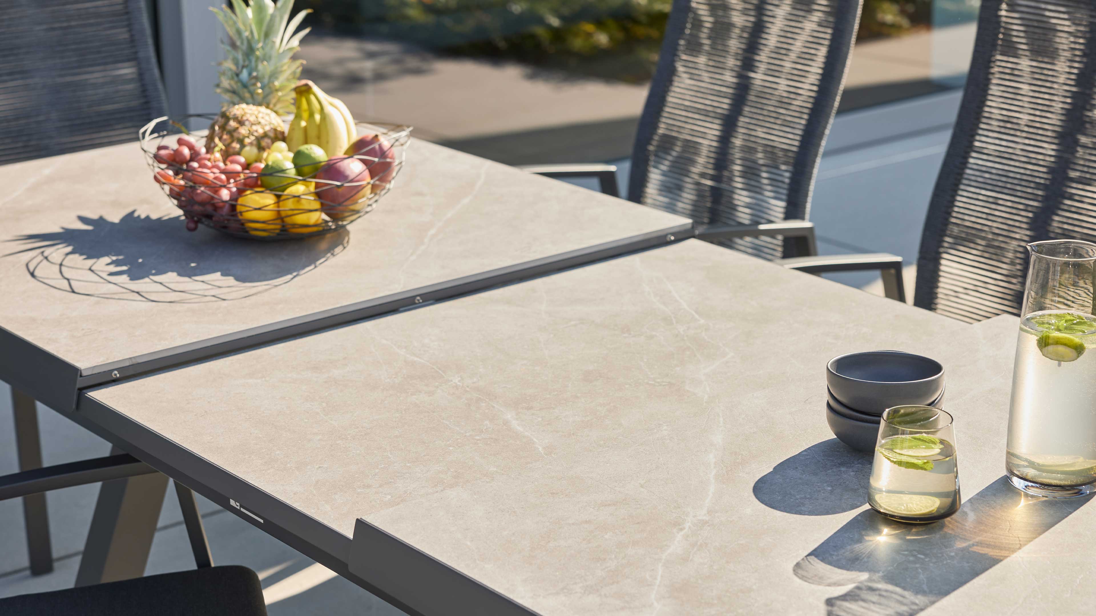 Rechteckiger Terrassentisch mit hellgrauer Steinoptikplatte und Metallrahmen, Obstkorb, Karaffe und Gläser.