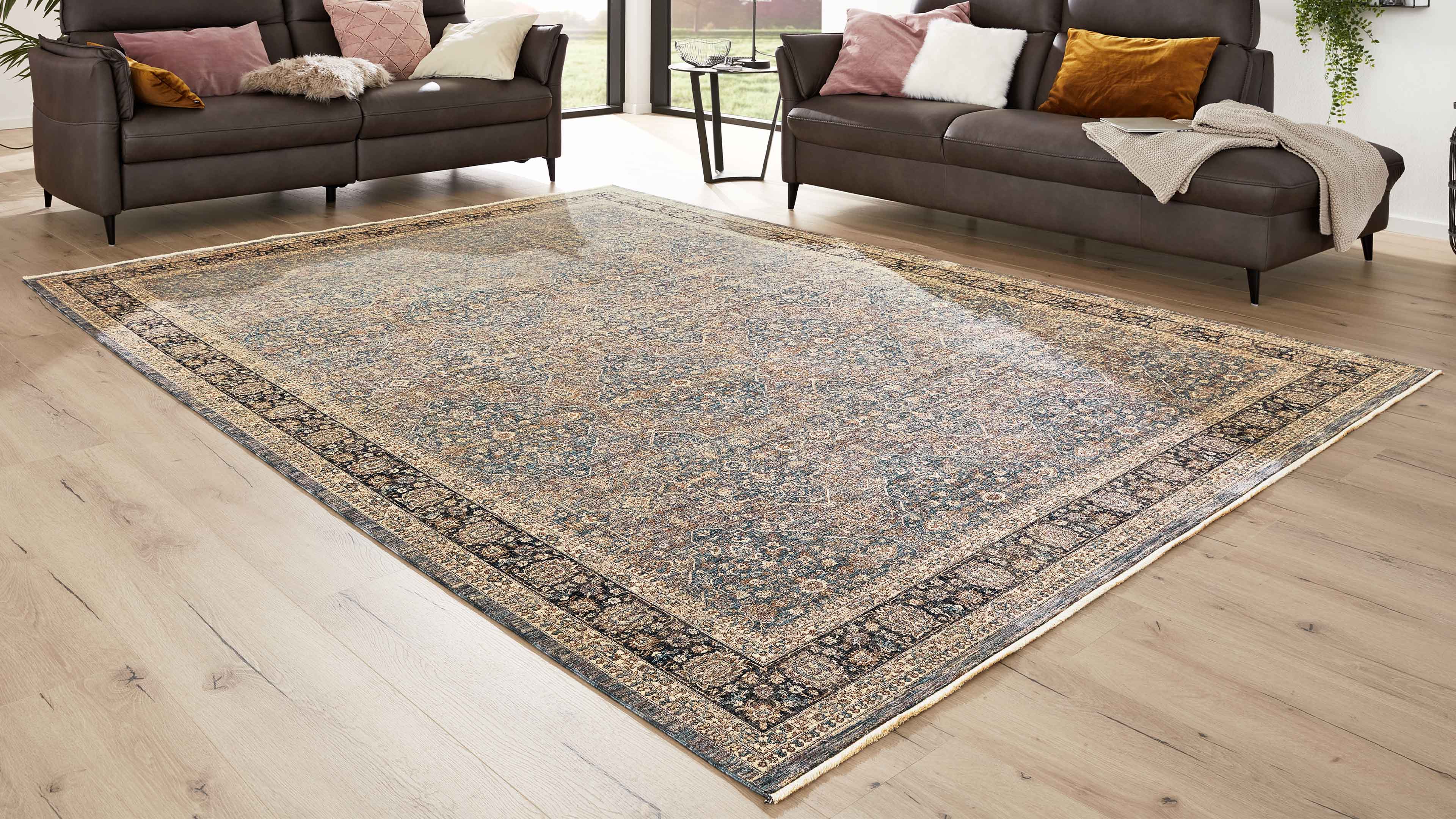 Großflächiger gemusterter Teppich auf hellem Holzfußboden, flankiert von zwei dunkelbraunen Sofas mit Kissen.