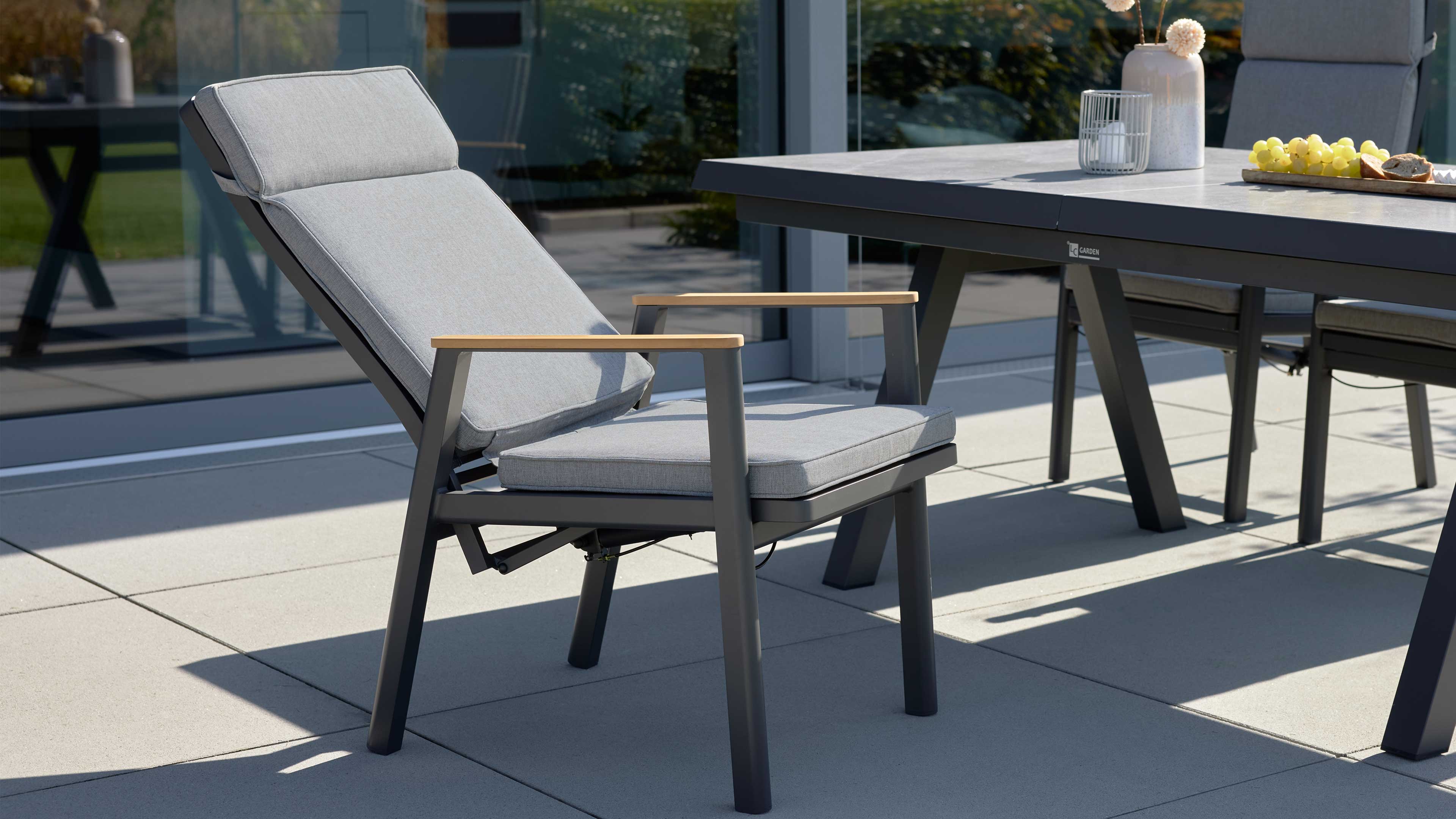 Hellgrau gepolsterter Sessel mit dunkelgrauem Metallgestell und Holzarmlehnen, neben einem dunklen Terrassentisch auf hellen Platten.