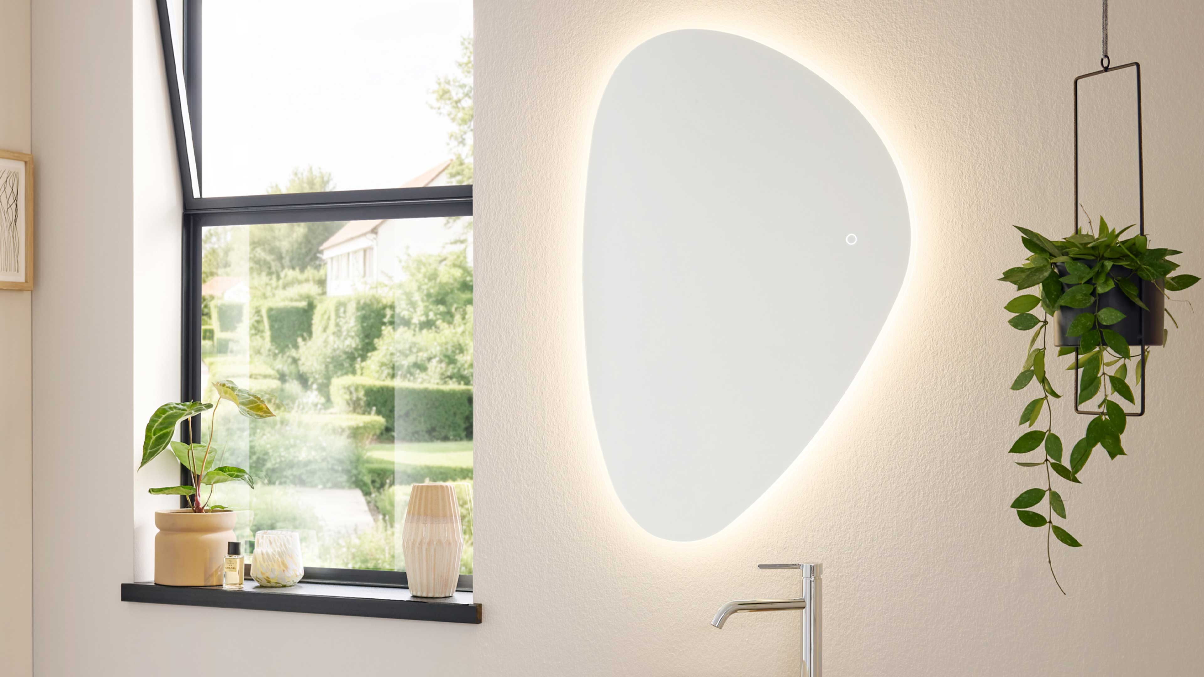 Großer unregelmäßiger, hinterleuchteter Wandspiegel über Waschbecken mit sichtbarer Armatur, Fensterbank mit Pflanzen und Vasen.