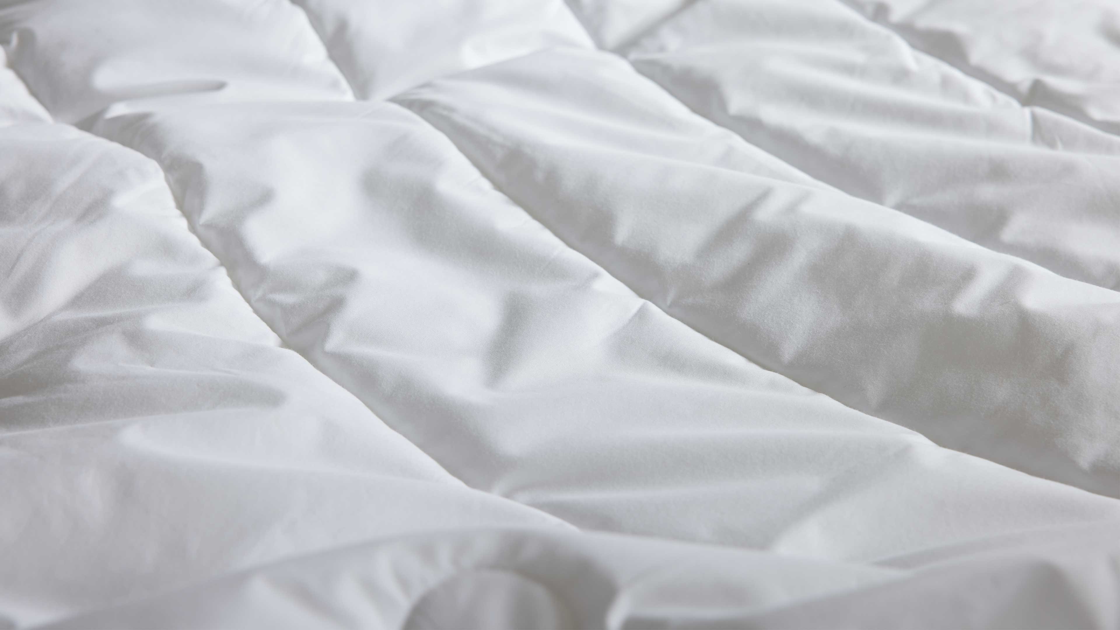 Weiße gesteppte Bettdecke mit parallelen, leicht gewölbten Kammernähten und weichen Falten in Nahaufnahme.