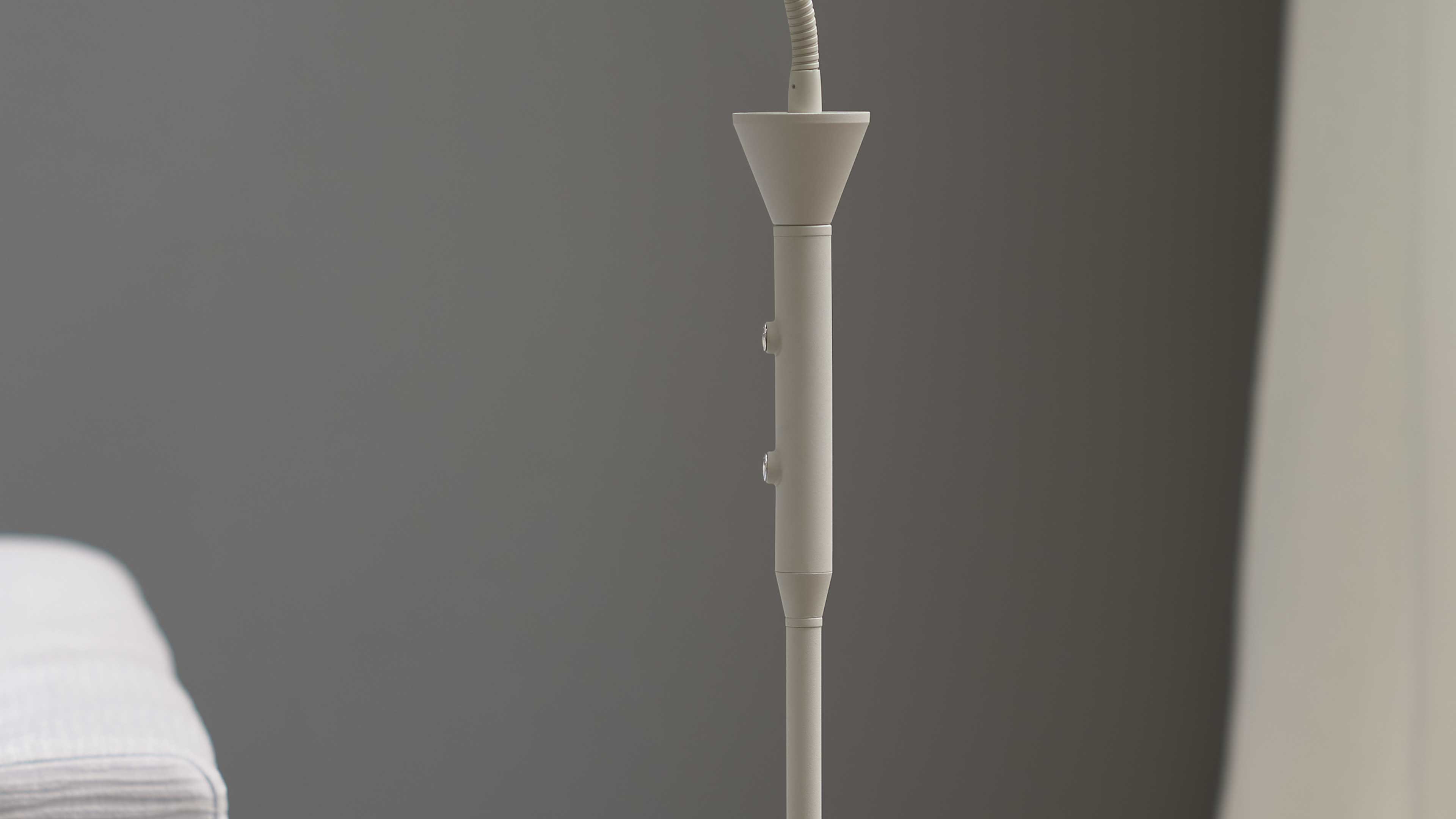 Schlanker cremefarbener Lampenstiel mit zwei runden Bedienknöpfen und Kabelansatz oben vor dunkelgrauem Hintergrund, links ein Stoffsofa.