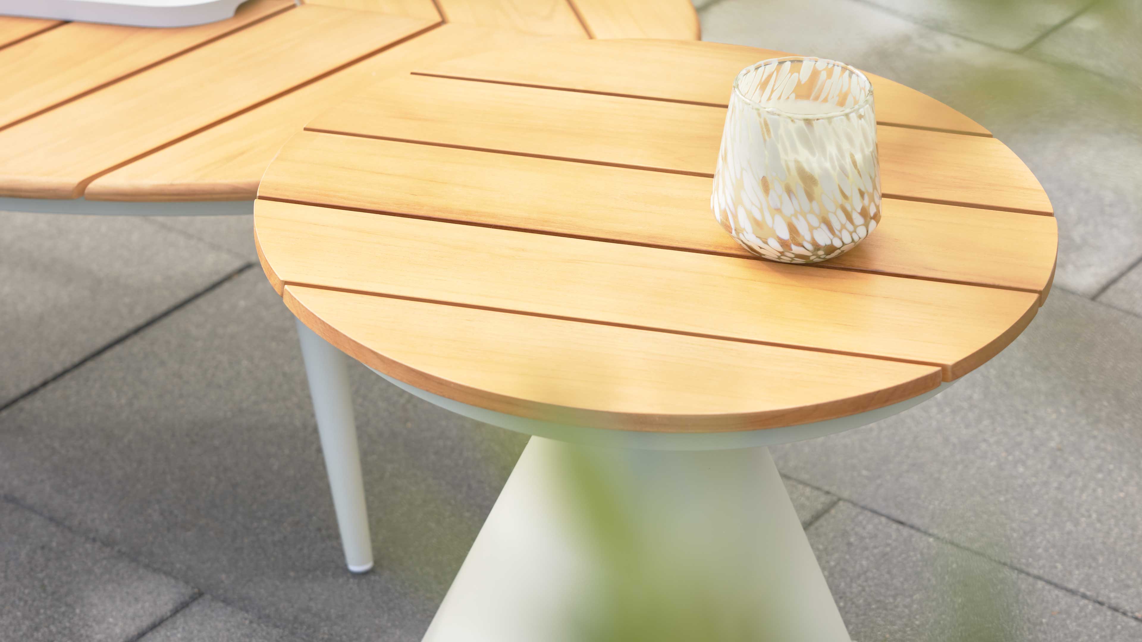 Runde Tischplatte mit hellen Holzlamellen auf weißem konischem Gestell, darauf eine weiß-beige gemusterte Vase, links Teil eines Tisches.