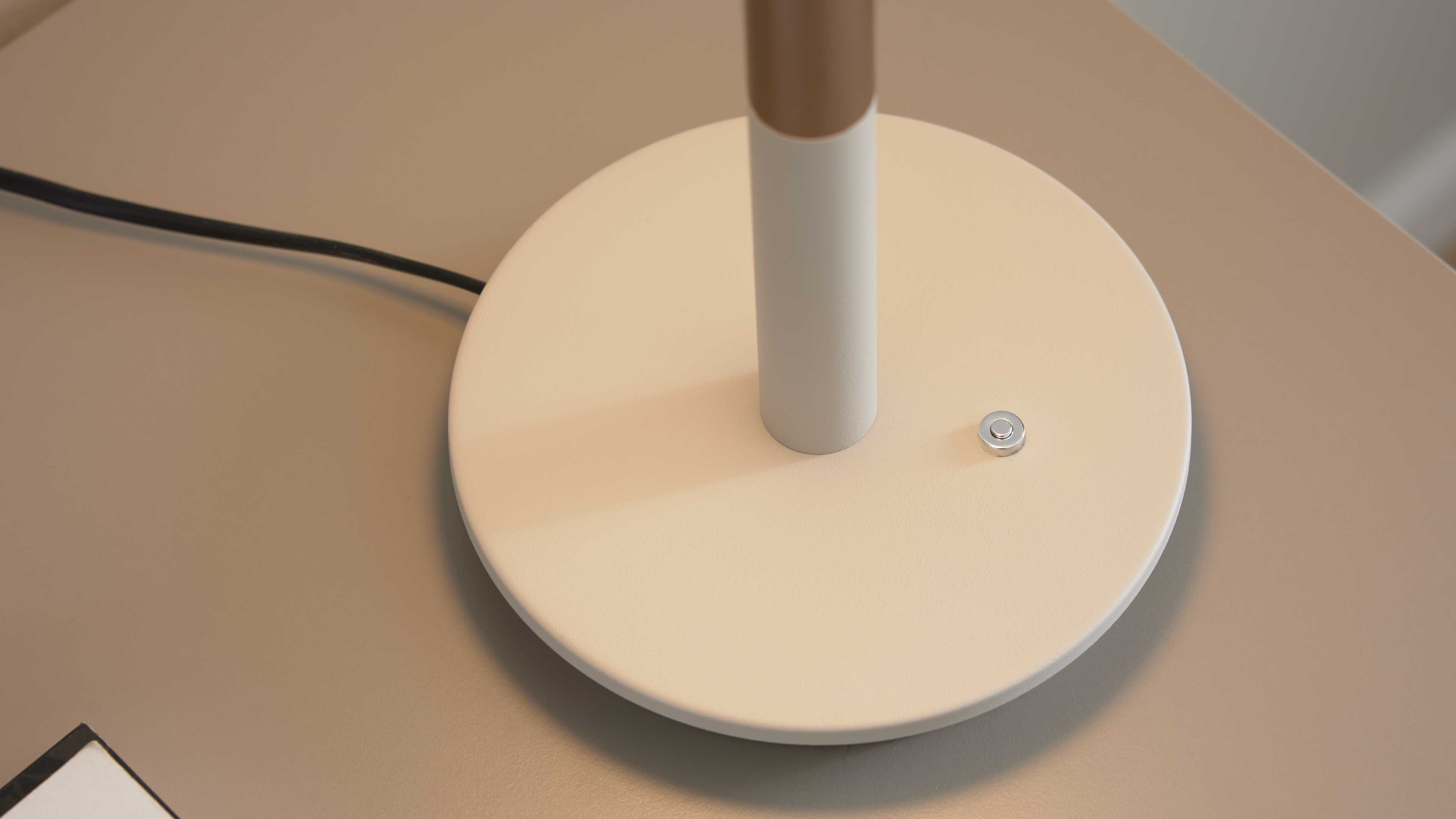 Runde helle Lampenfußplatte mit mittigem zylindrischem Stiel, kleinem silbernem Druckknopf und schwarzem Kabel auf beigem Tisch.
