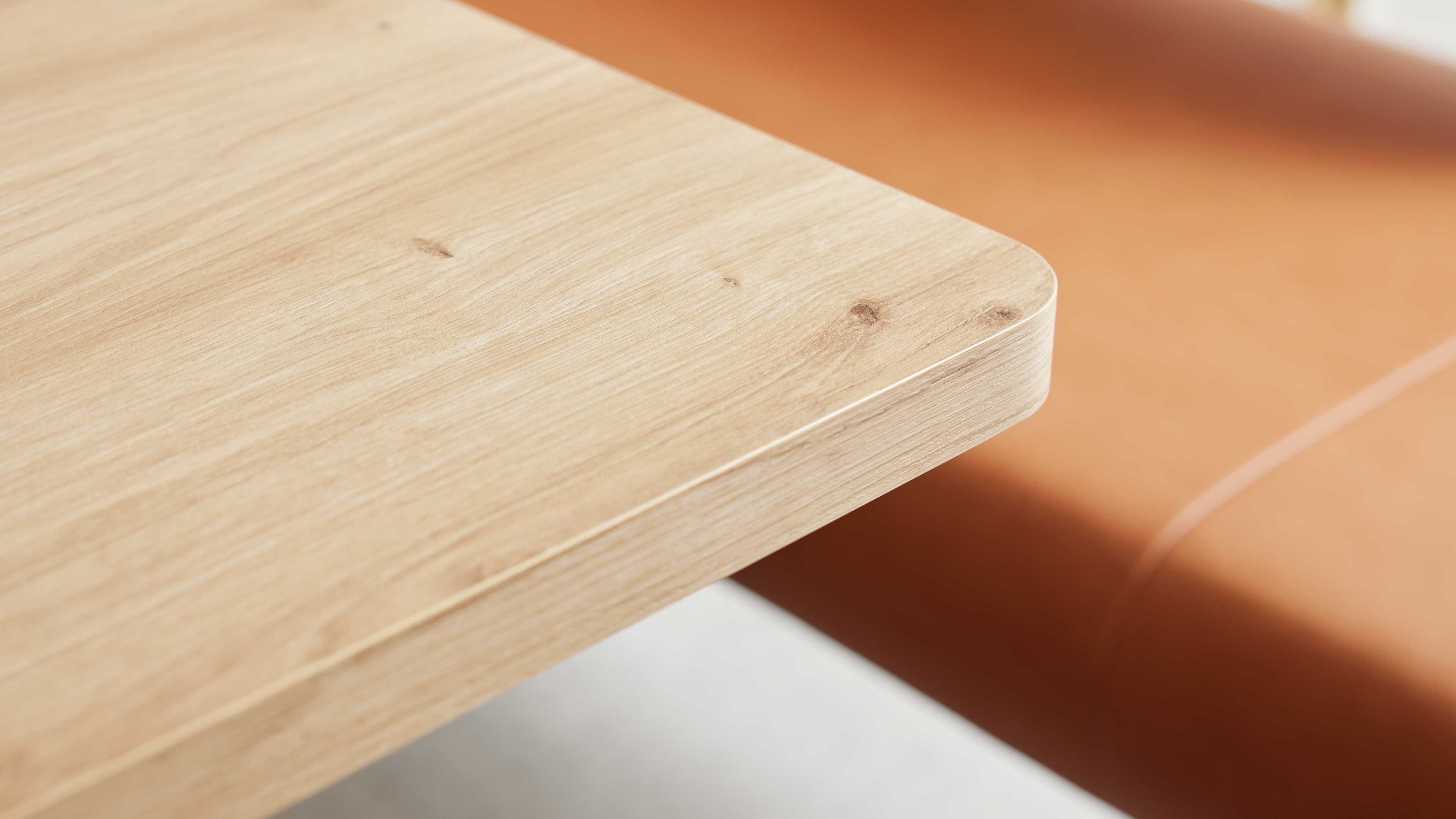 Nahaufnahme einer abgerundeten Tischplatte aus hellem Holz mit sichtbarer Maserung und Knoten, vor orangebrauner Polsterfläche.