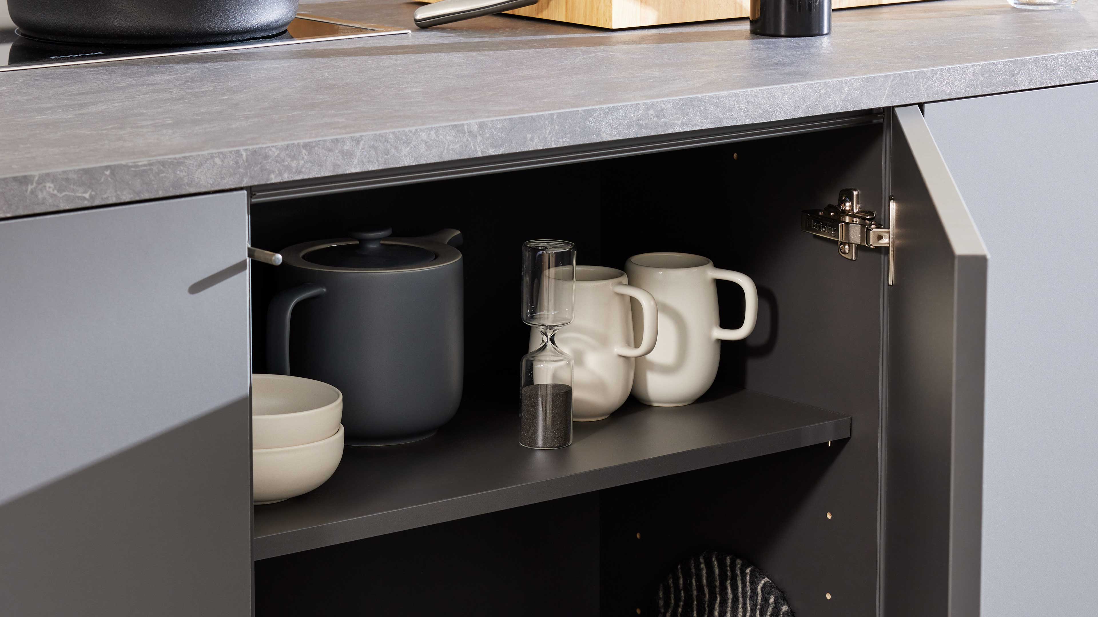 Offenes graues Küchenschrankfach mit grauer Teekanne, zwei übereinander gestapelten Schalen, Sanduhr und zwei weißen Tassen auf der Ablage.