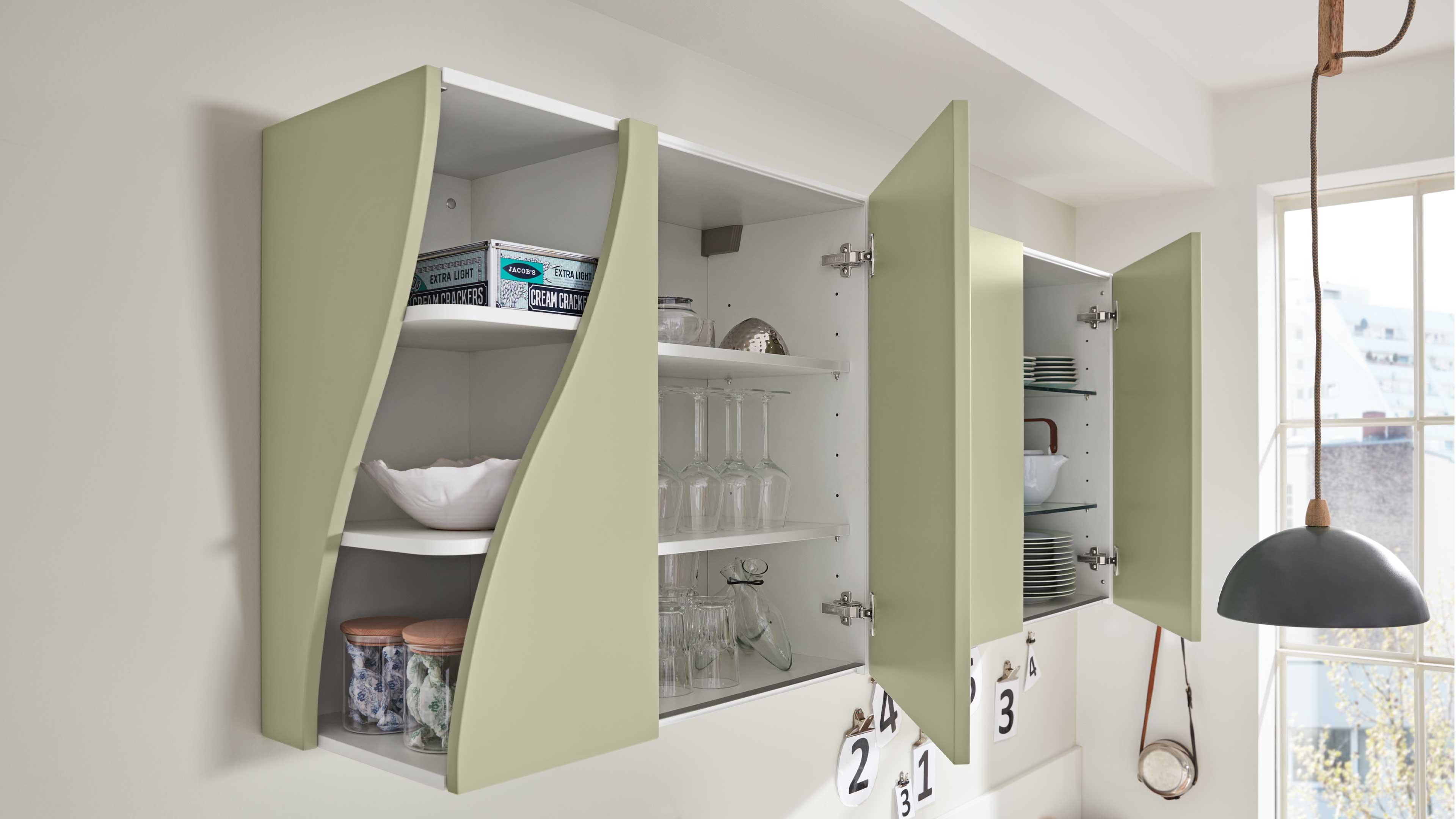 Dreiteilige, wandhängende Küchenschränke mit matten hellgrünen, geschwungenen Türen, weißem Innenraum und eingeräumten Gläsern und Tellern.