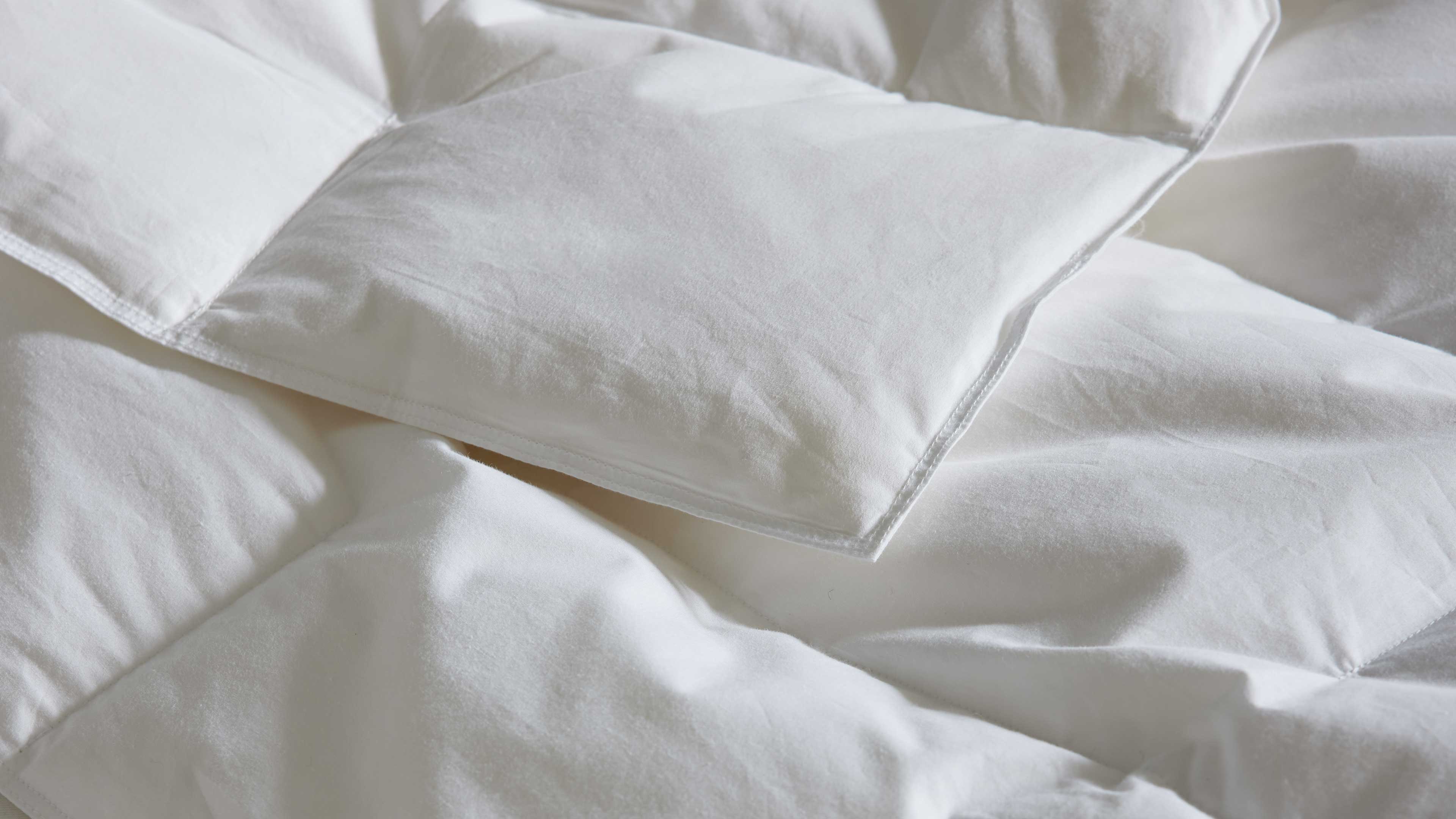 Weiße gesteppte Bettdecke in Nahaufnahme mit quadratischen Steppfeldern, sichtbaren Nähten und weichen, leicht gewellten Falten.