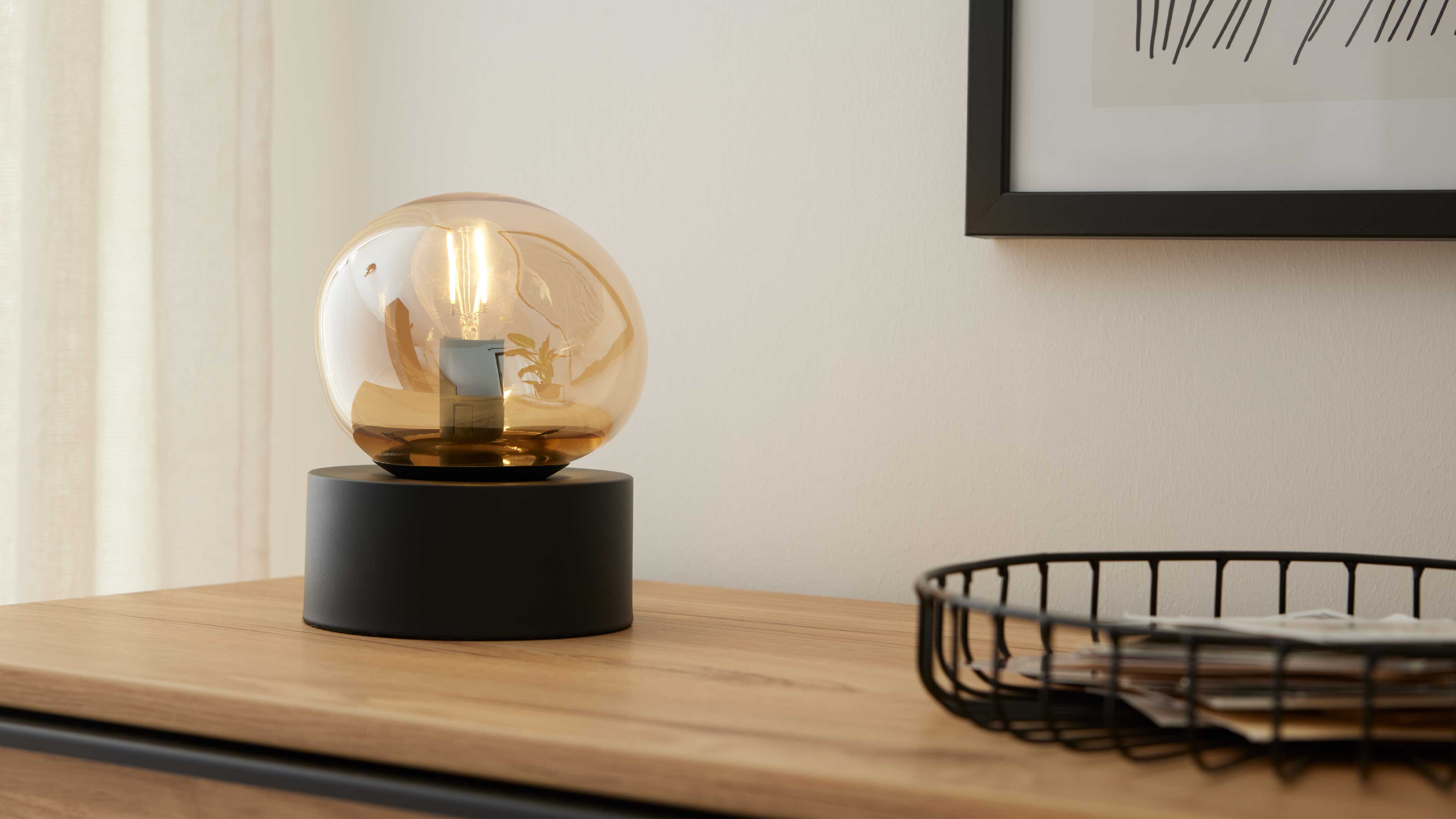 Tischlampe mit bernsteinfarbener Glasglocke auf schwarzem Zylinderfuß auf Holzkommode, rechts Drahtkorb mit Papieren.