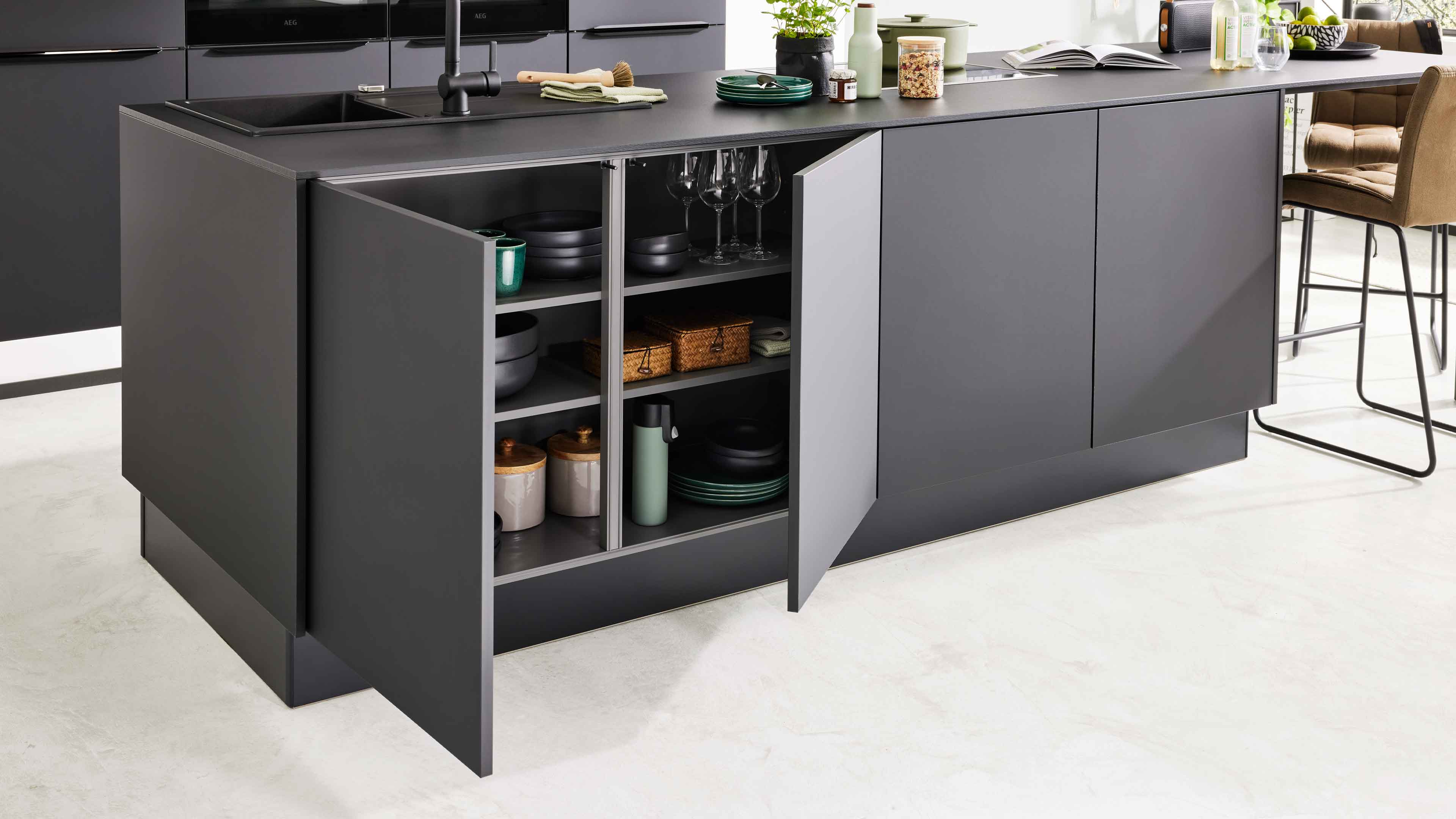 Dunkelgraue Kücheninsel mit offenen Schranktüren, sichtbaren Regalen mit Geschirr, Gläsern, Dosen, Spüle oben links und Barhocker rechts.