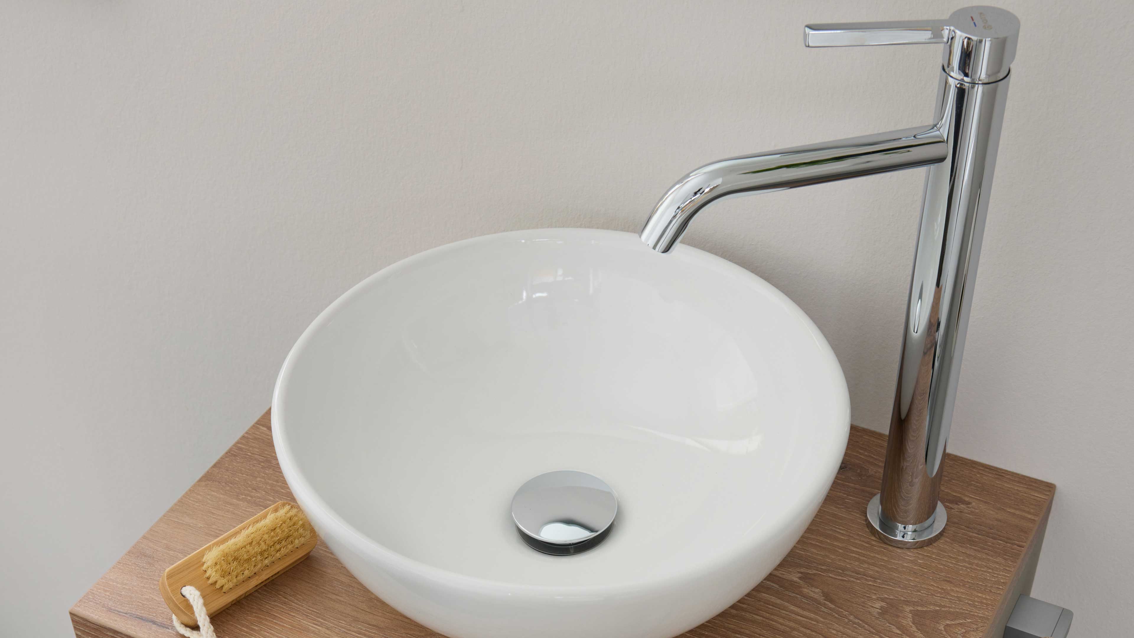 Weißes rundes Aufsatzbecken auf Holzplatte mit rundem Chromablauf, hoher verchromter Einhebelarmatur rechts und kleiner Bürste links.
