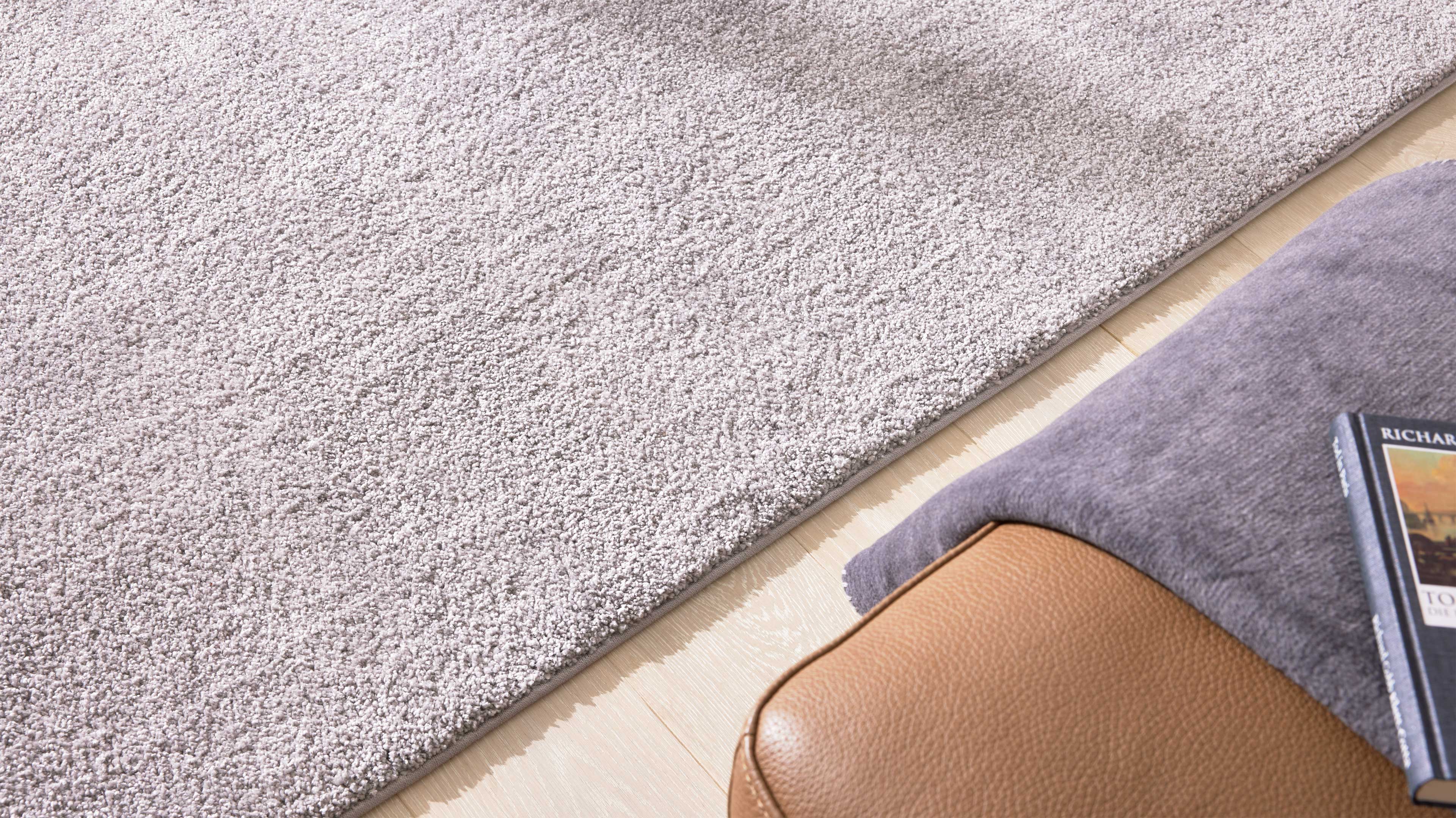 Großflächiger, hellgrauer dichtfloriger Teppich; rechts unten braunes Lederhocker-Eck mit grauer Decke und Buch.