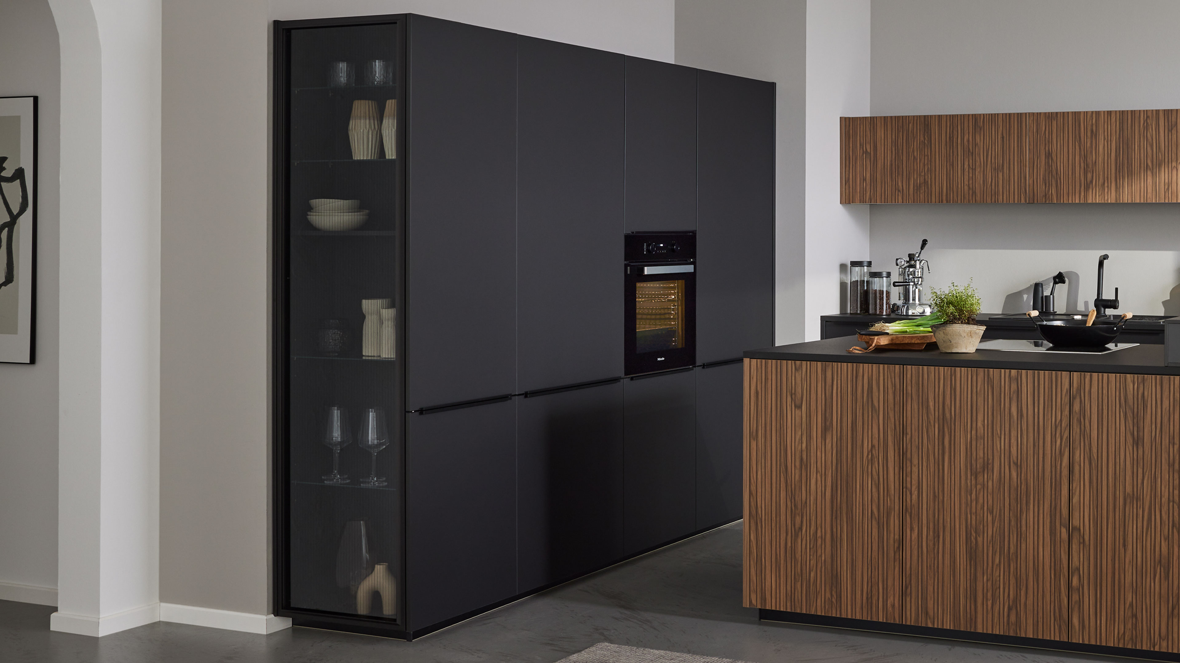 Hoher, mattschwarzer Küchenblock mit Glastür und eingebautem Backofen, daneben Holzfront-Insel und hängender Holzschrank.