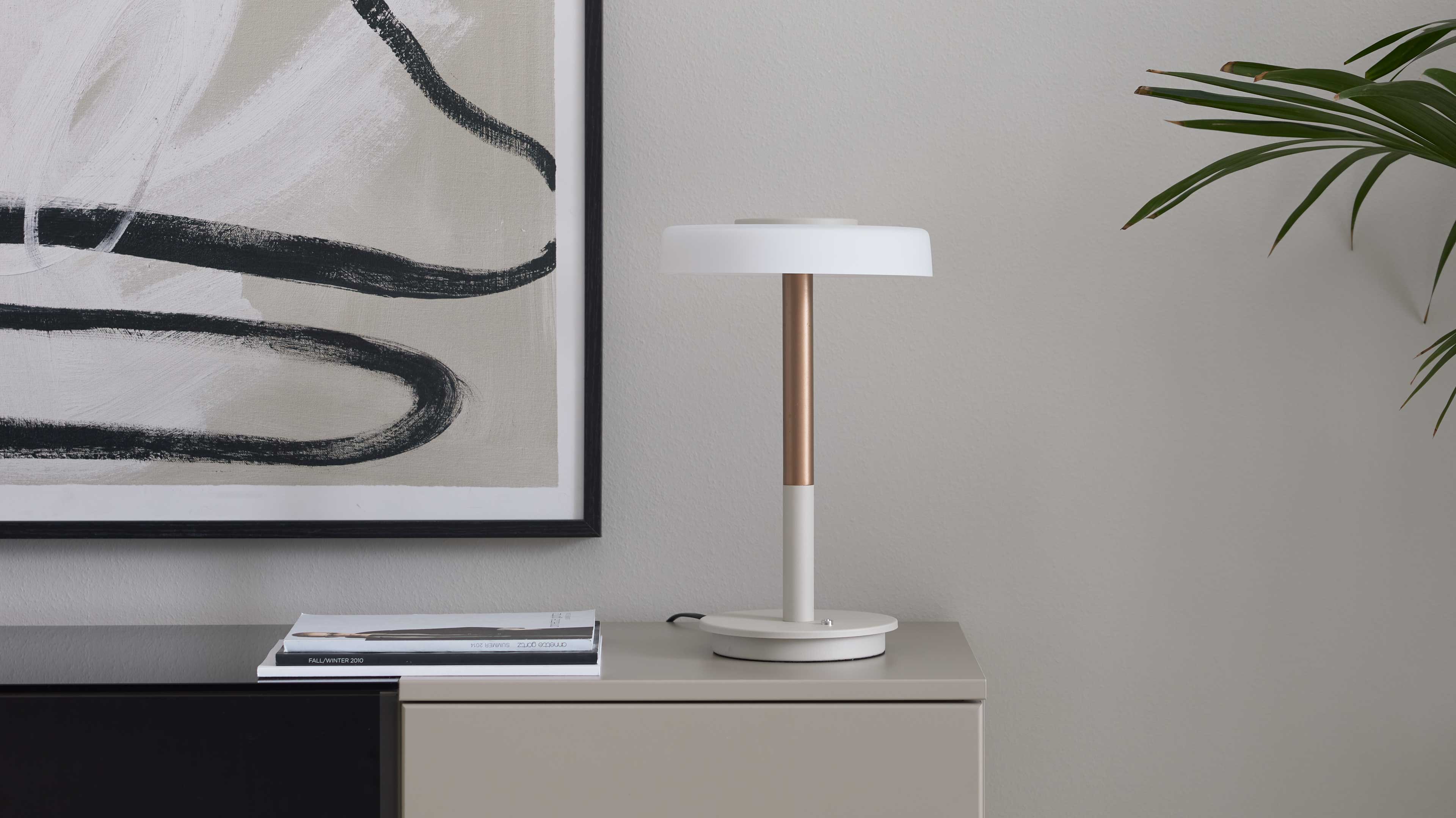 Tischlampe mit weißem Rundschirm und kupferfarbenem Schaft auf beigem Sideboard; links schwarz gerahmtes Bild, Magazin und Pflanzblätter rechts.