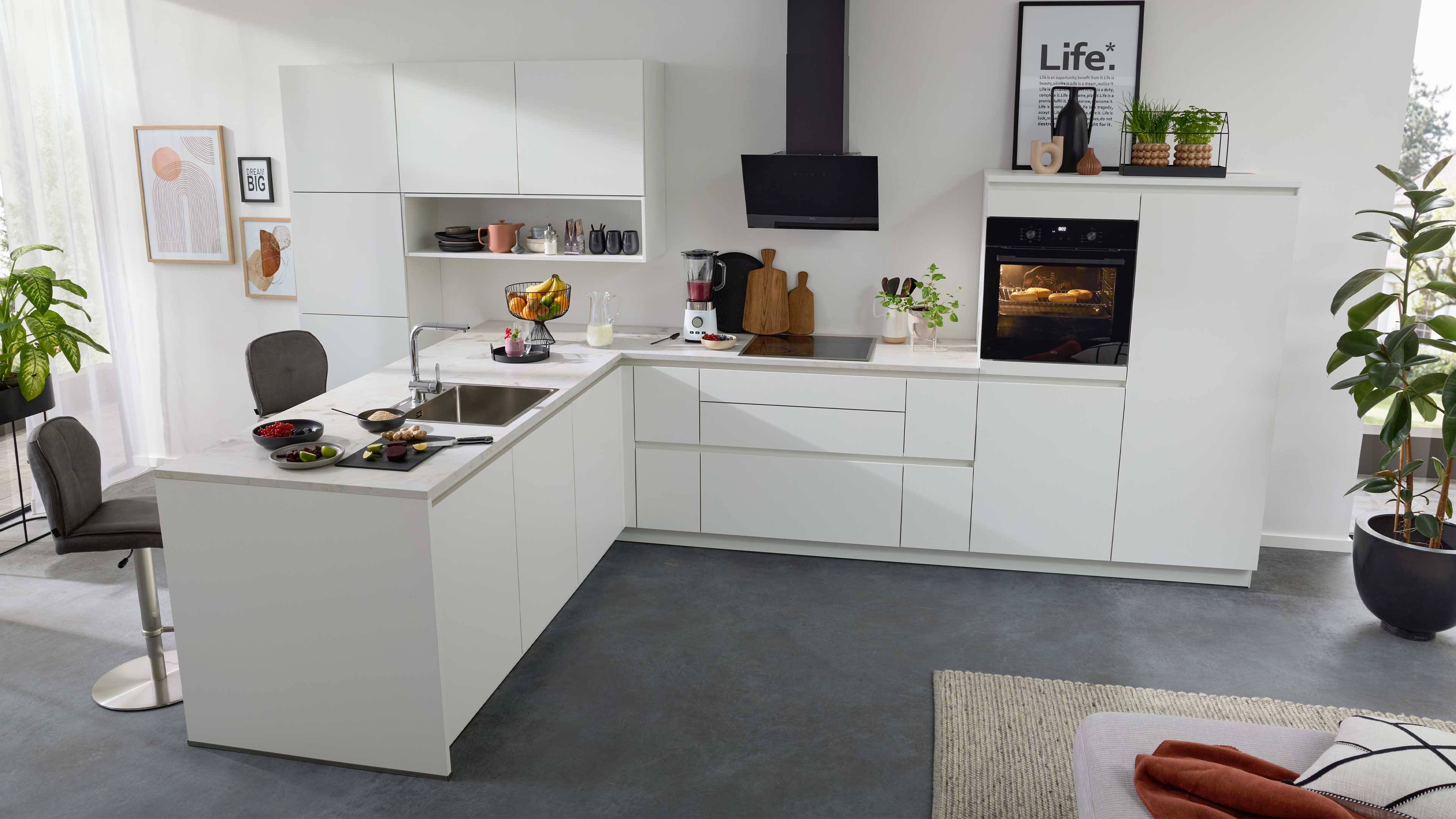 Weiße L‑Küche mit weißer Arbeitsplatte, Edelstahlspüle, Kochfeld, eingebautem Backofen mit Sicht auf Gebäck, Obstkorb und Pflanzen.